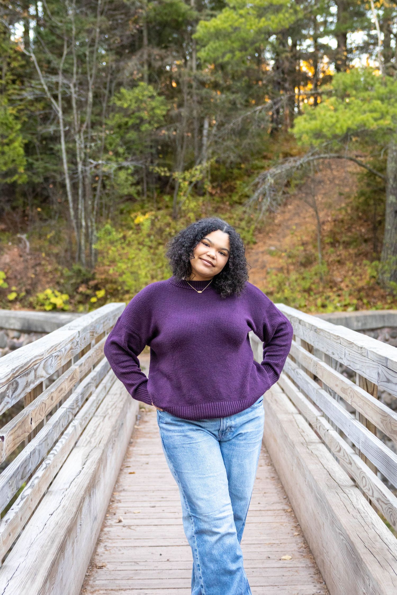 Person in purple sweater and jeans poses playfully on wooden bridge surrounded by autumn trees.