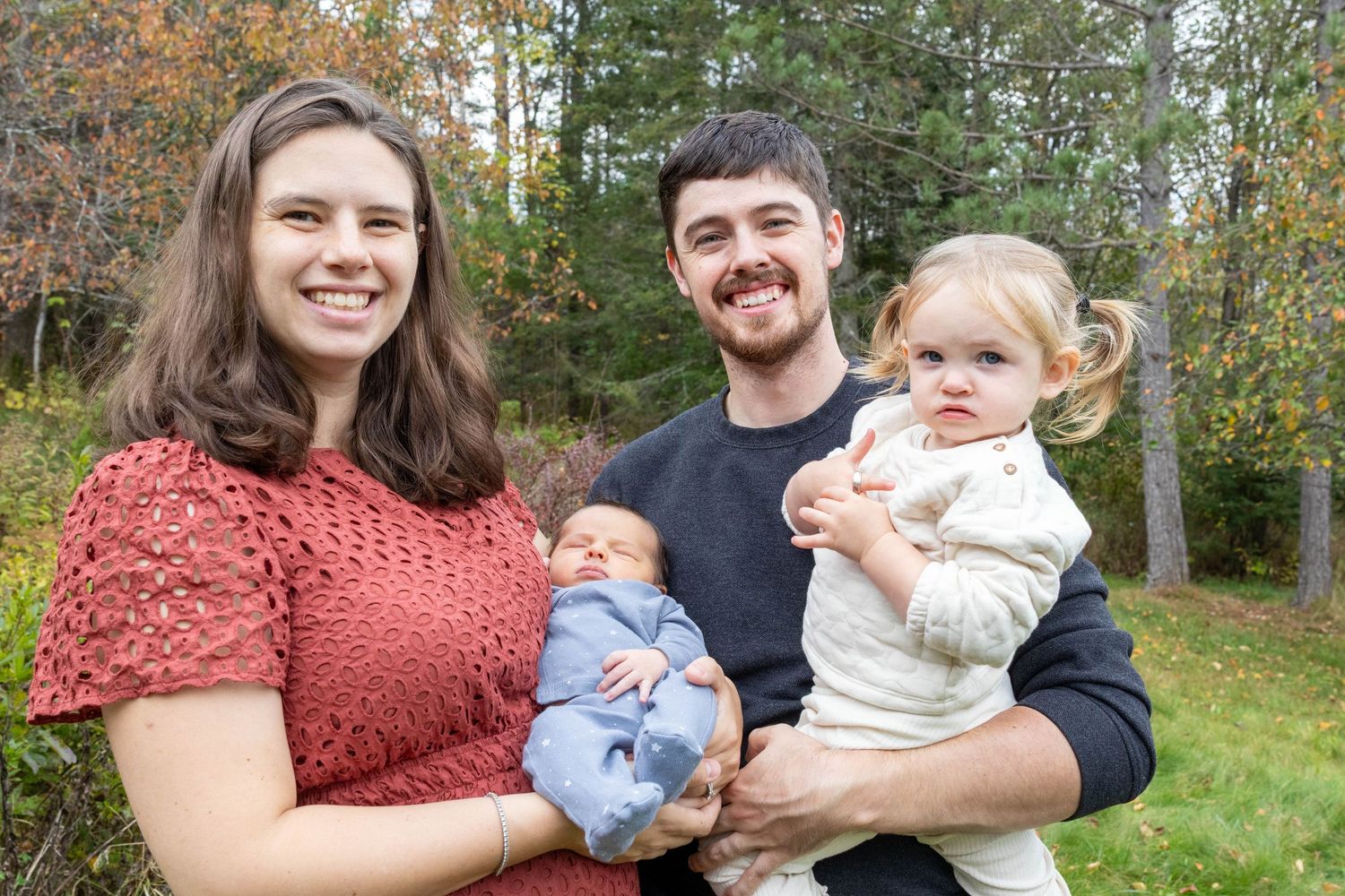 A family sitting together outdoors in autumn with trees and fallen leaves visible in the background.