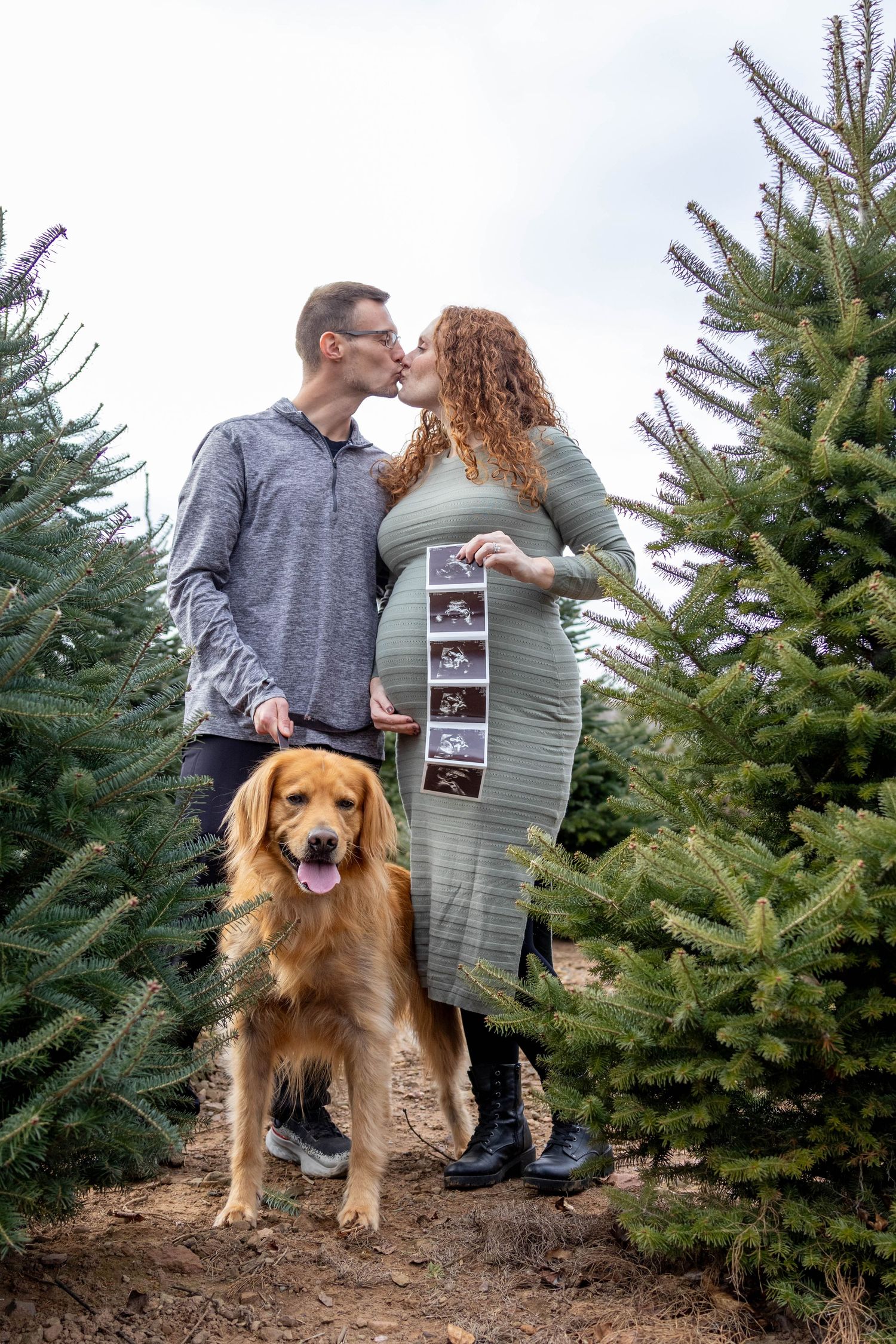 A couple shares a tender moment at a Christmas tree farm while their golden retriever sits happily nearby.