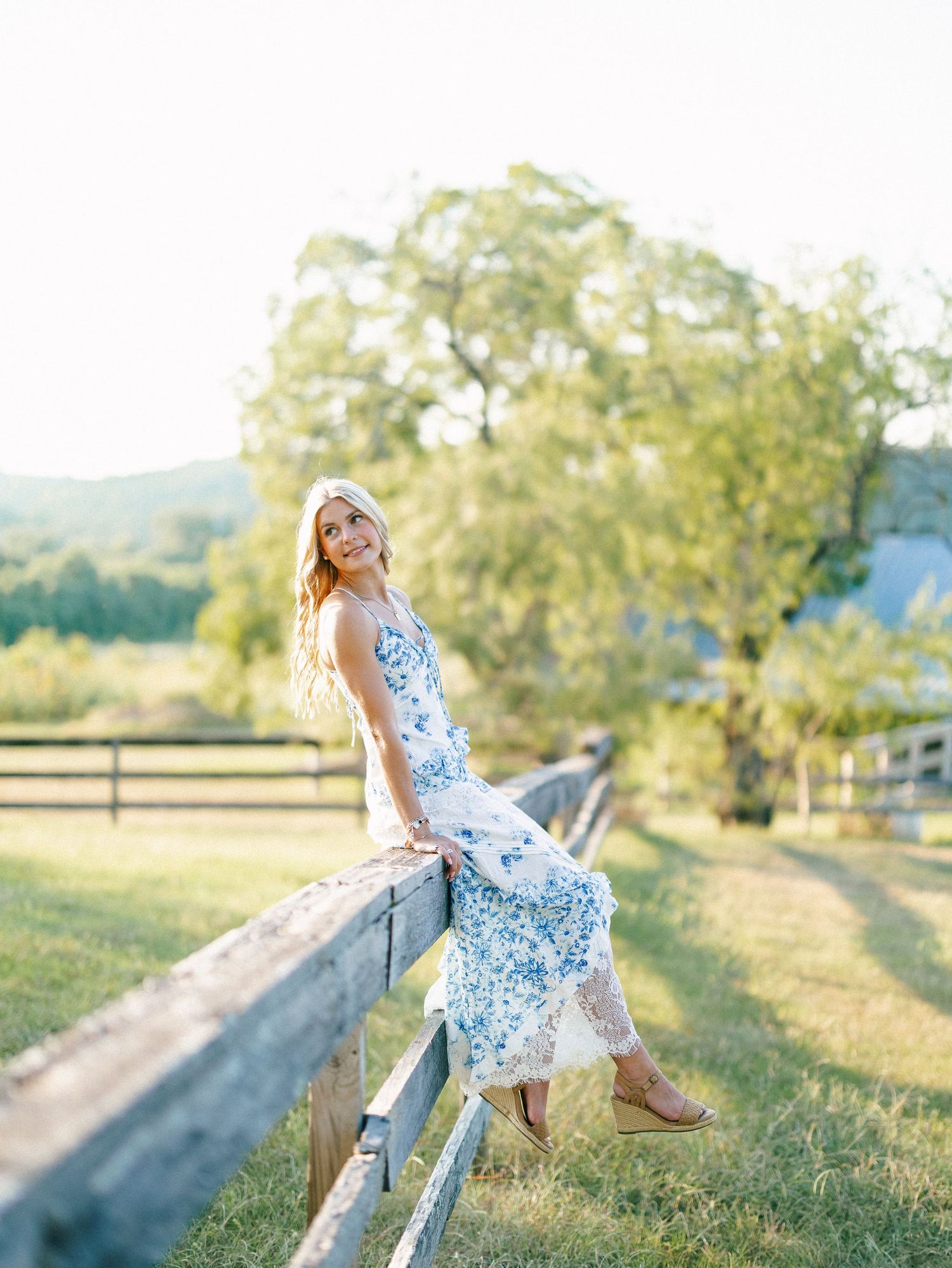 Woman in blue floral maxi dress leaning on rustic wooden fence at golden hour on a scenic farm.