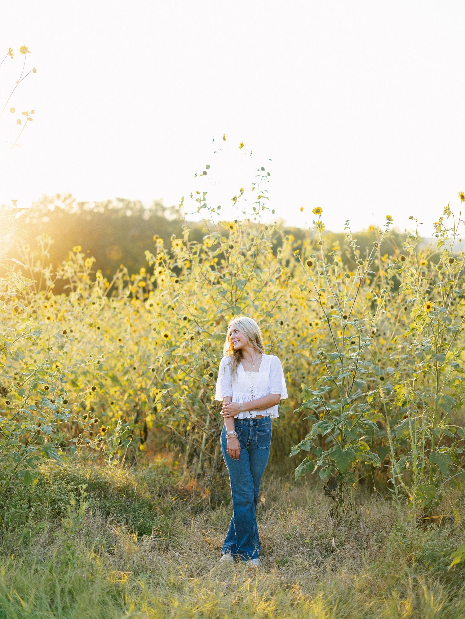 Woman in white top and jeans standing in a sunflower field bathed in golden hour sunlight.
