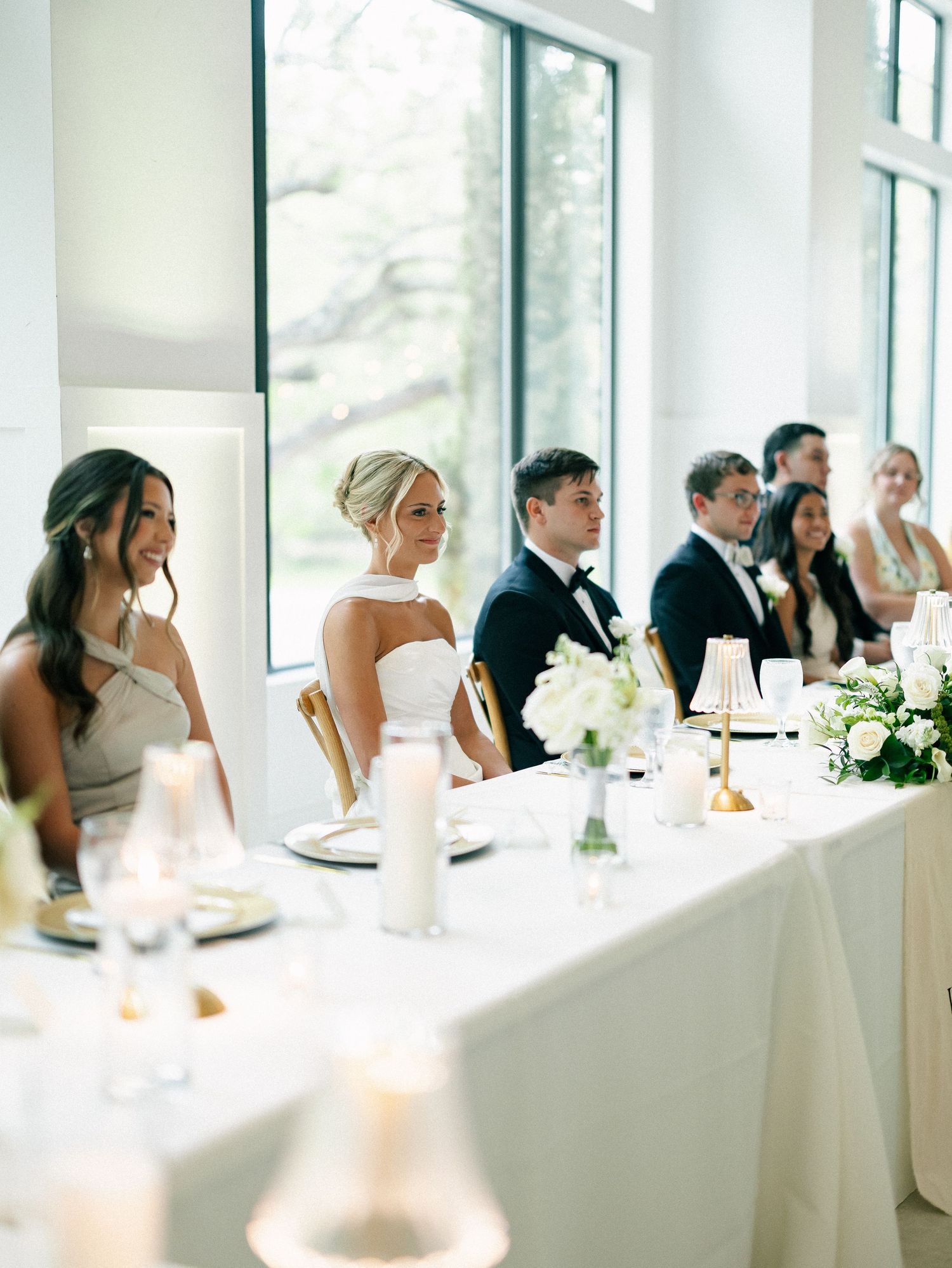 Wedding party seated at elegant white head table with candles and floral arrangements during reception.