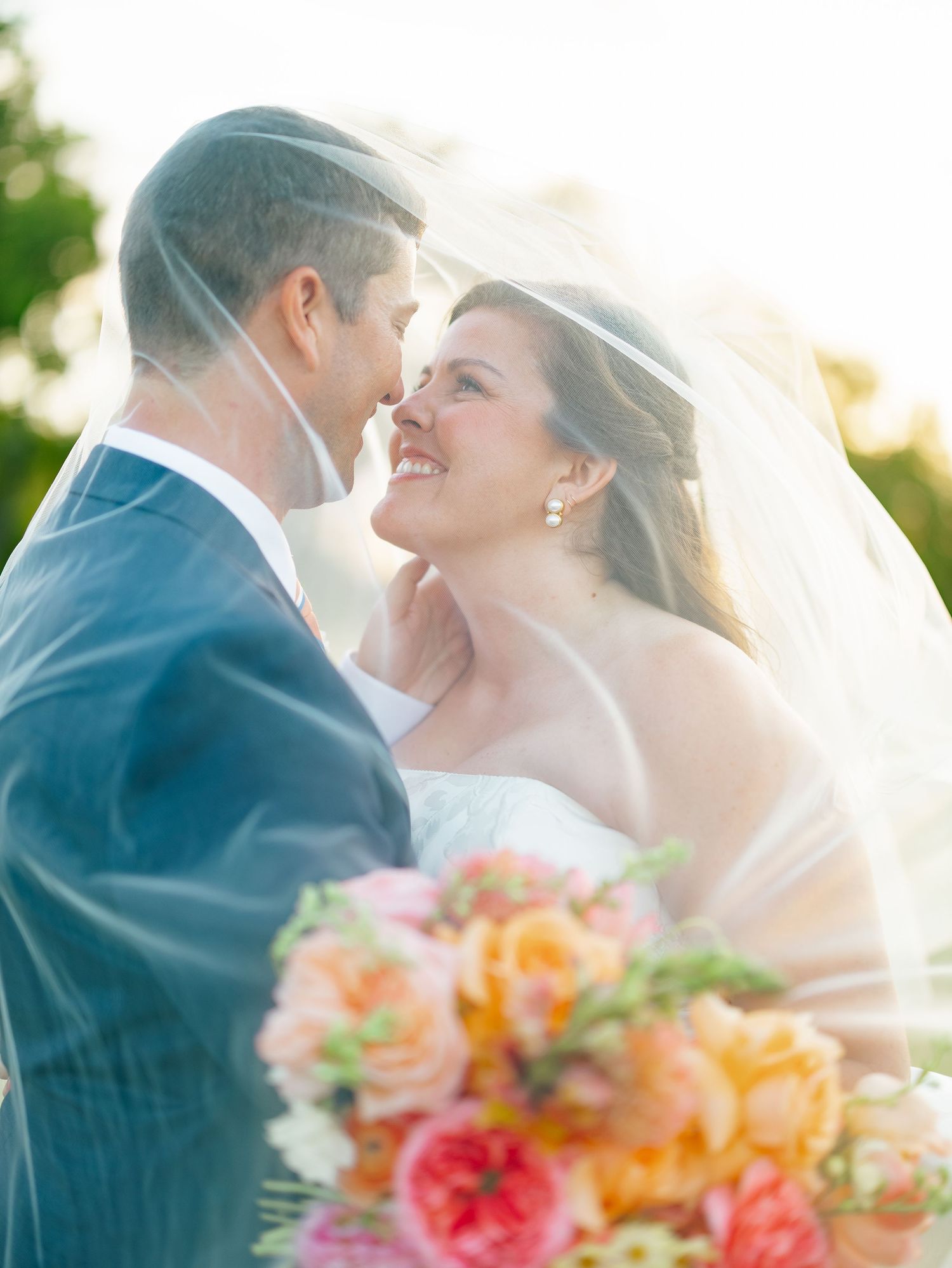 Bride and groom sharing romantic moment under veil with colorful peach and pink bridal bouquet.