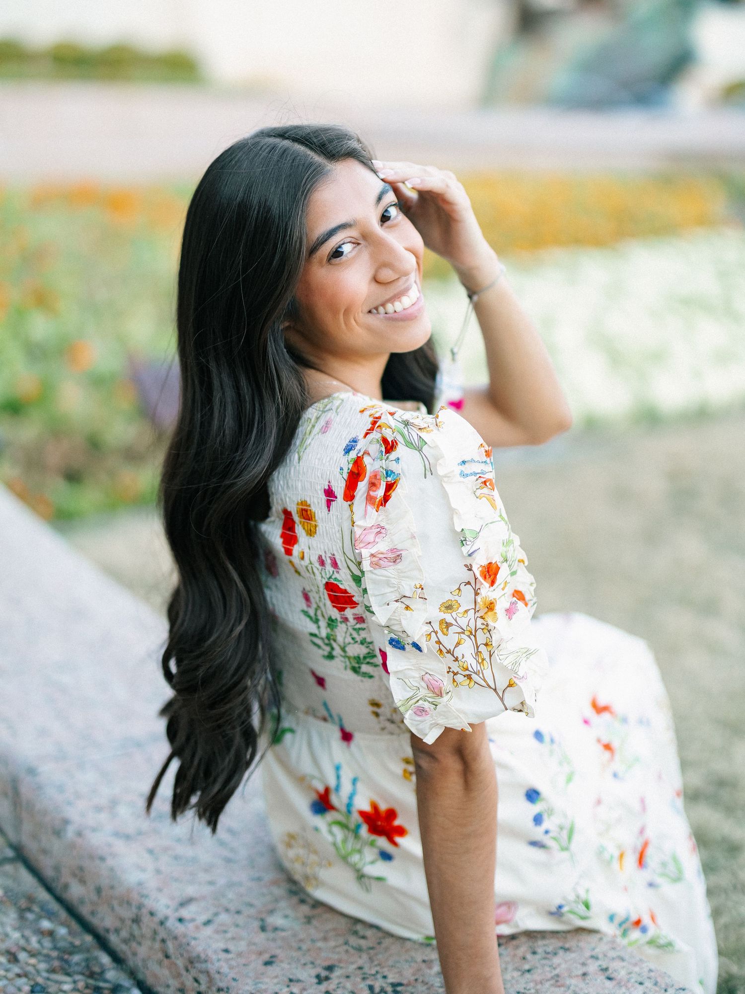 Young woman in floral white dress smiling over shoulder, long dark hair, seated outdoors with colorful flowers behind.
