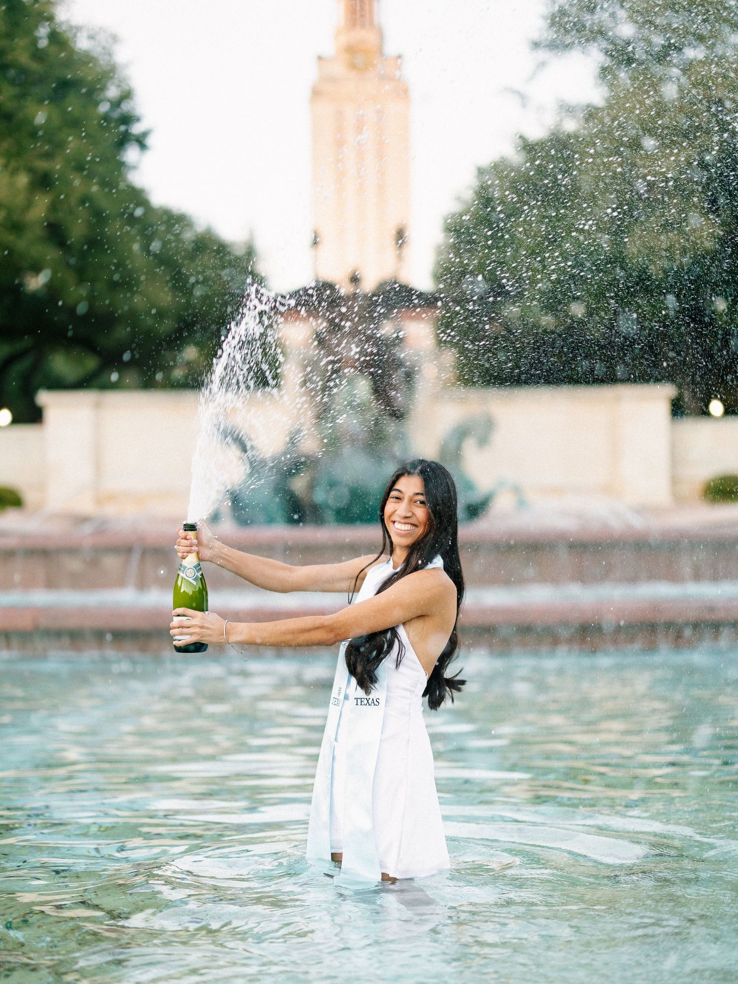 Woman in white dress celebrating in fountain, popping champagne bottle with university tower in background.