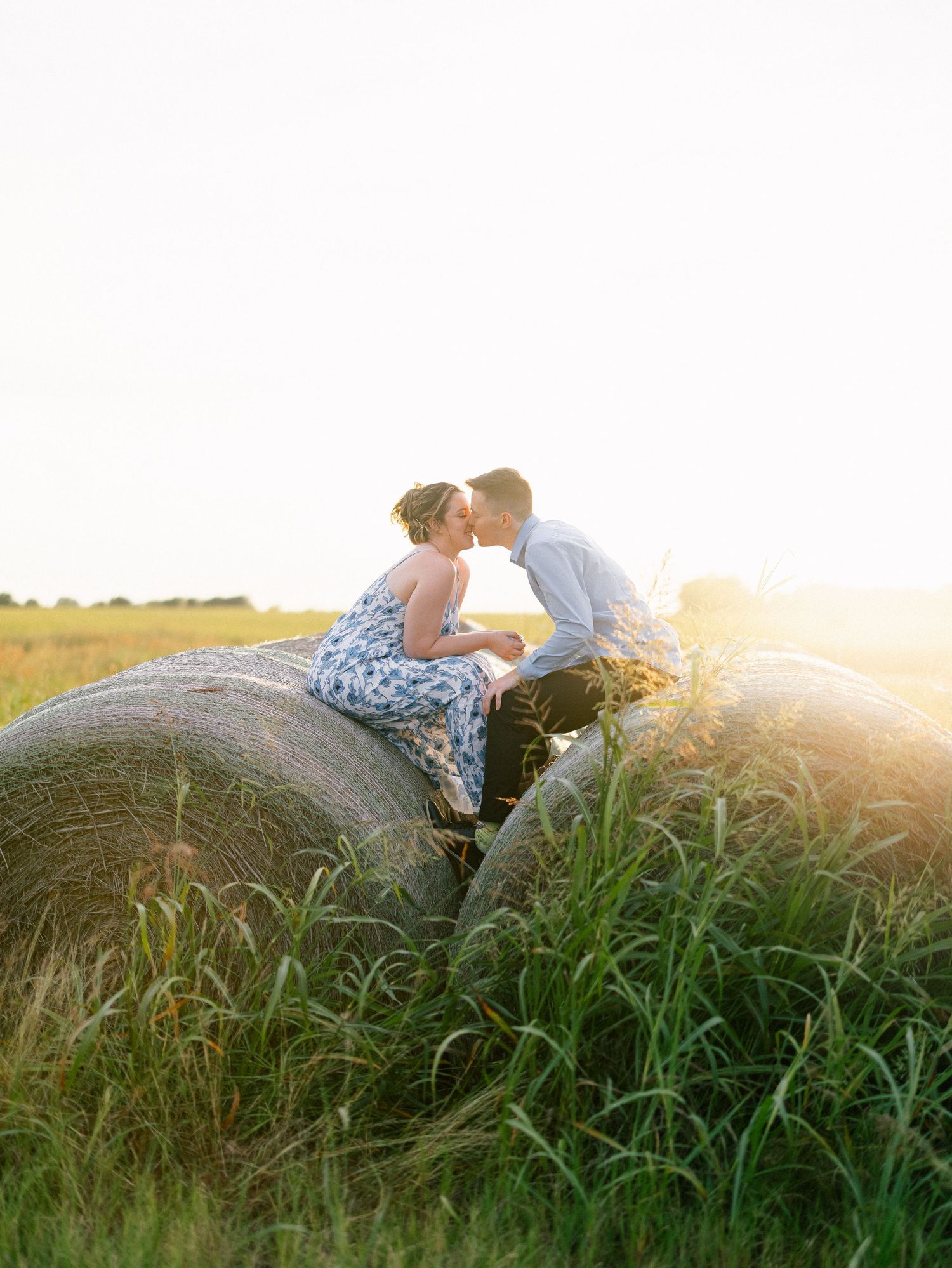 Couple sharing a tender kiss on a hay bale in a sunlit field during a romantic engagement session.