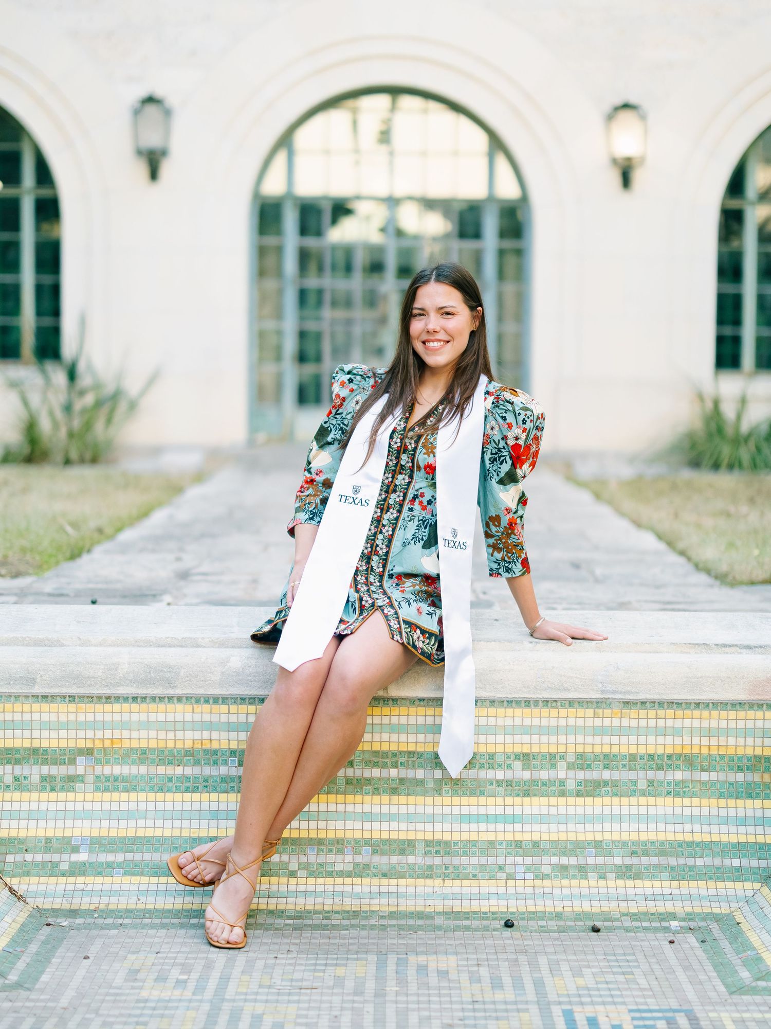Smiling graduate in floral dress and white sash sits on mosaic steps outside elegant arched building.