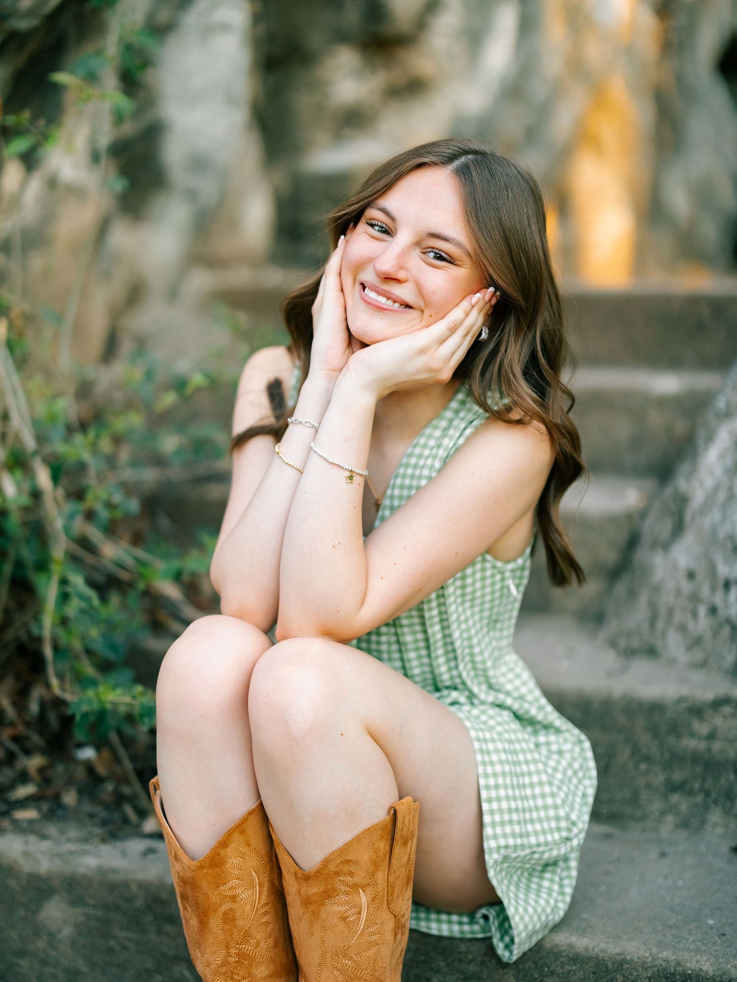 Smiling young woman in green gingham dress and cowboy boots sitting on stone steps with hands on cheeks.