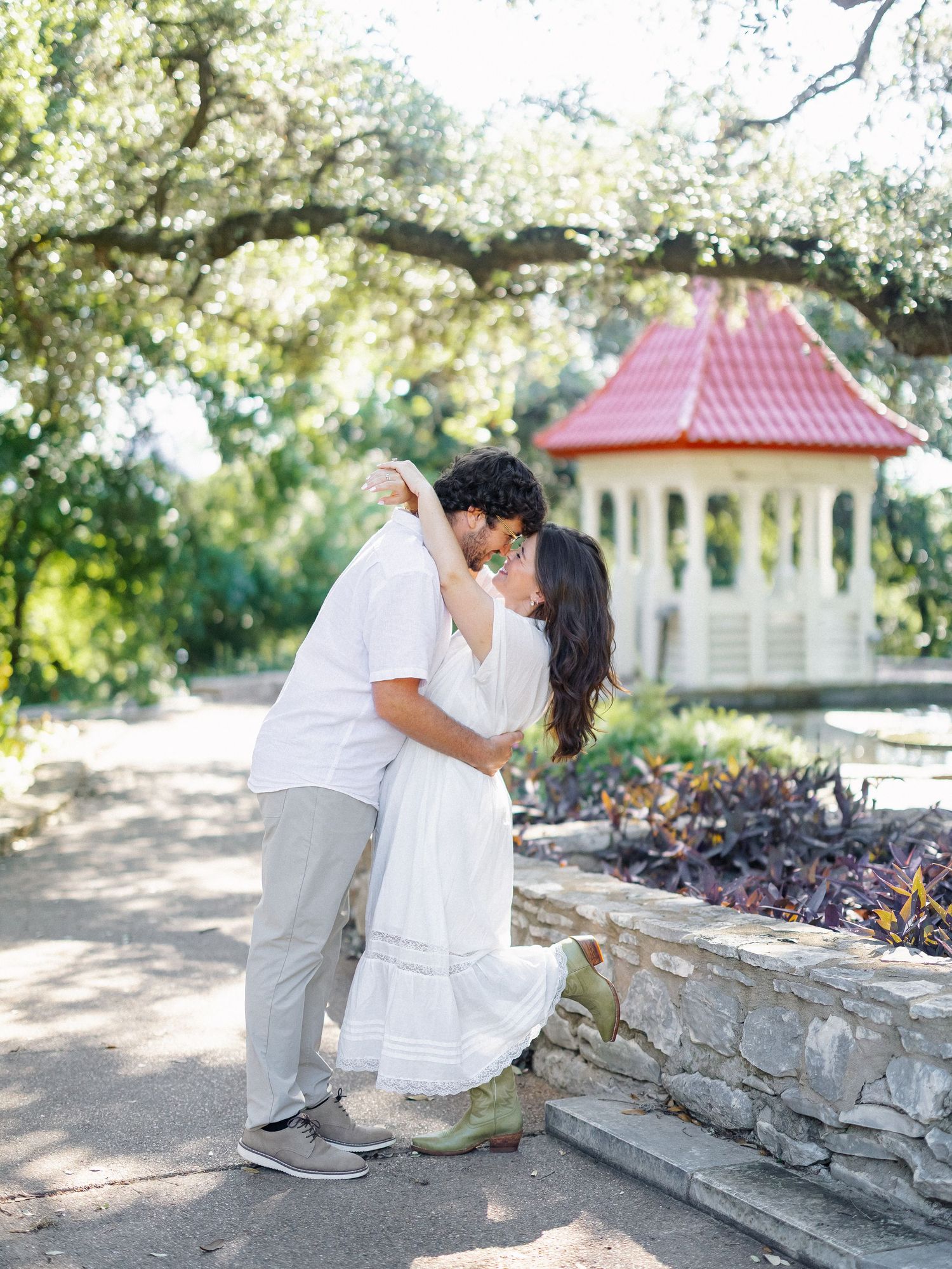 Couple sharing a romantic dip kiss near a stone wall and red-roofed gazebo surrounded by lush greenery.