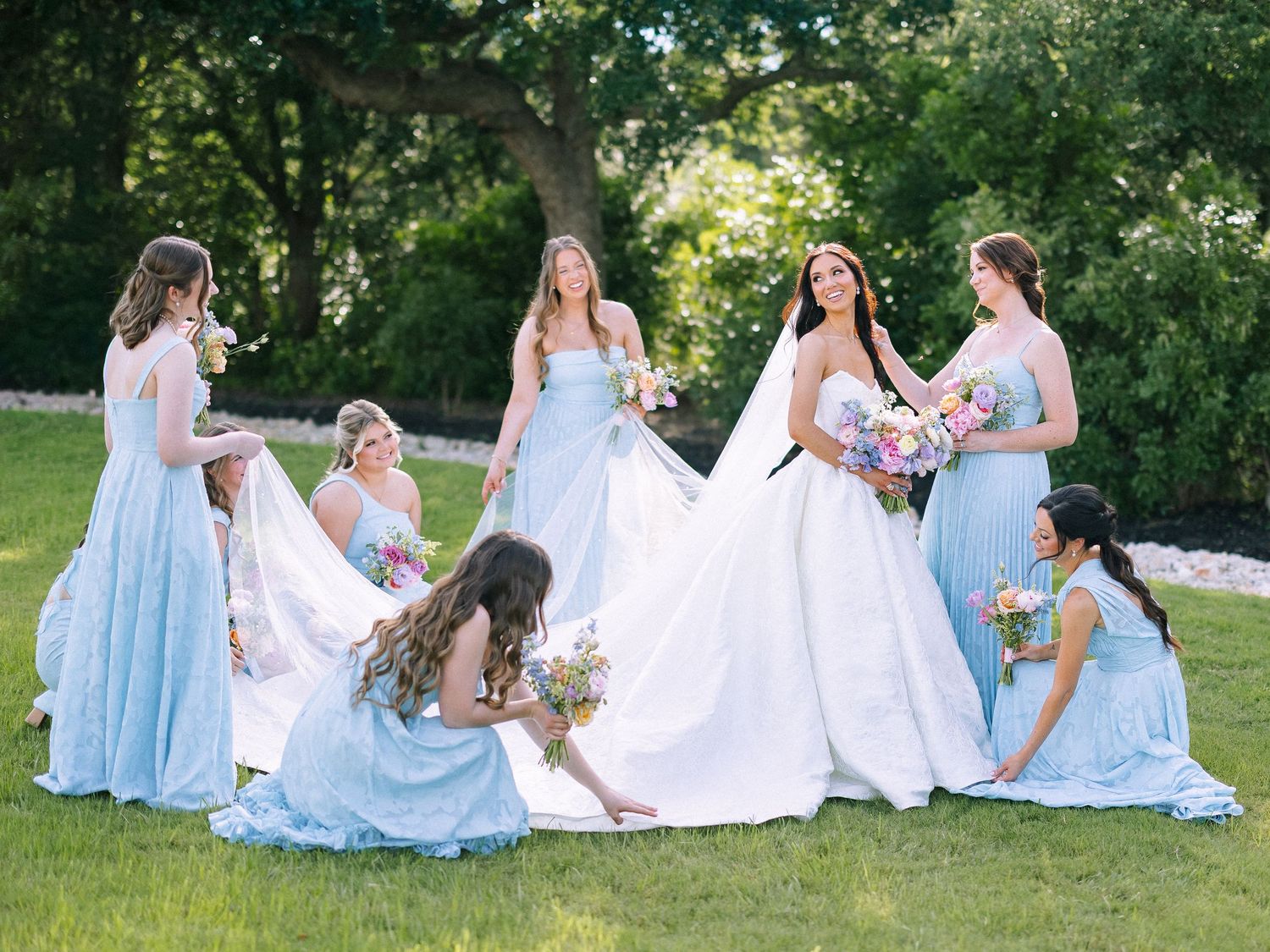 Bride in white gown surrounded by bridesmaids in light blue dresses arranging her train on green lawn.
