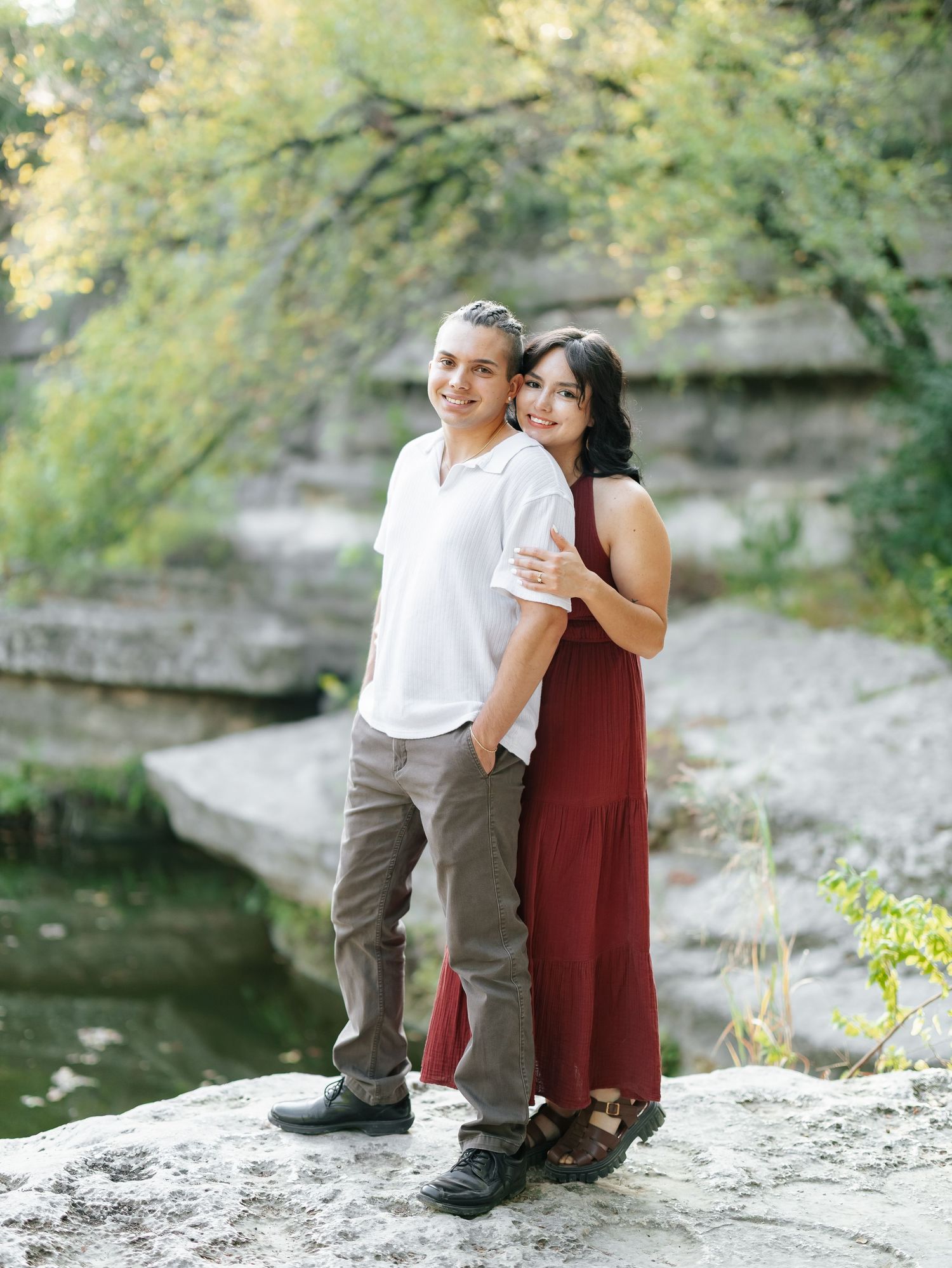 Couple posing on rocks near a creek, woman in burgundy dress embracing man in white shirt outdoors.