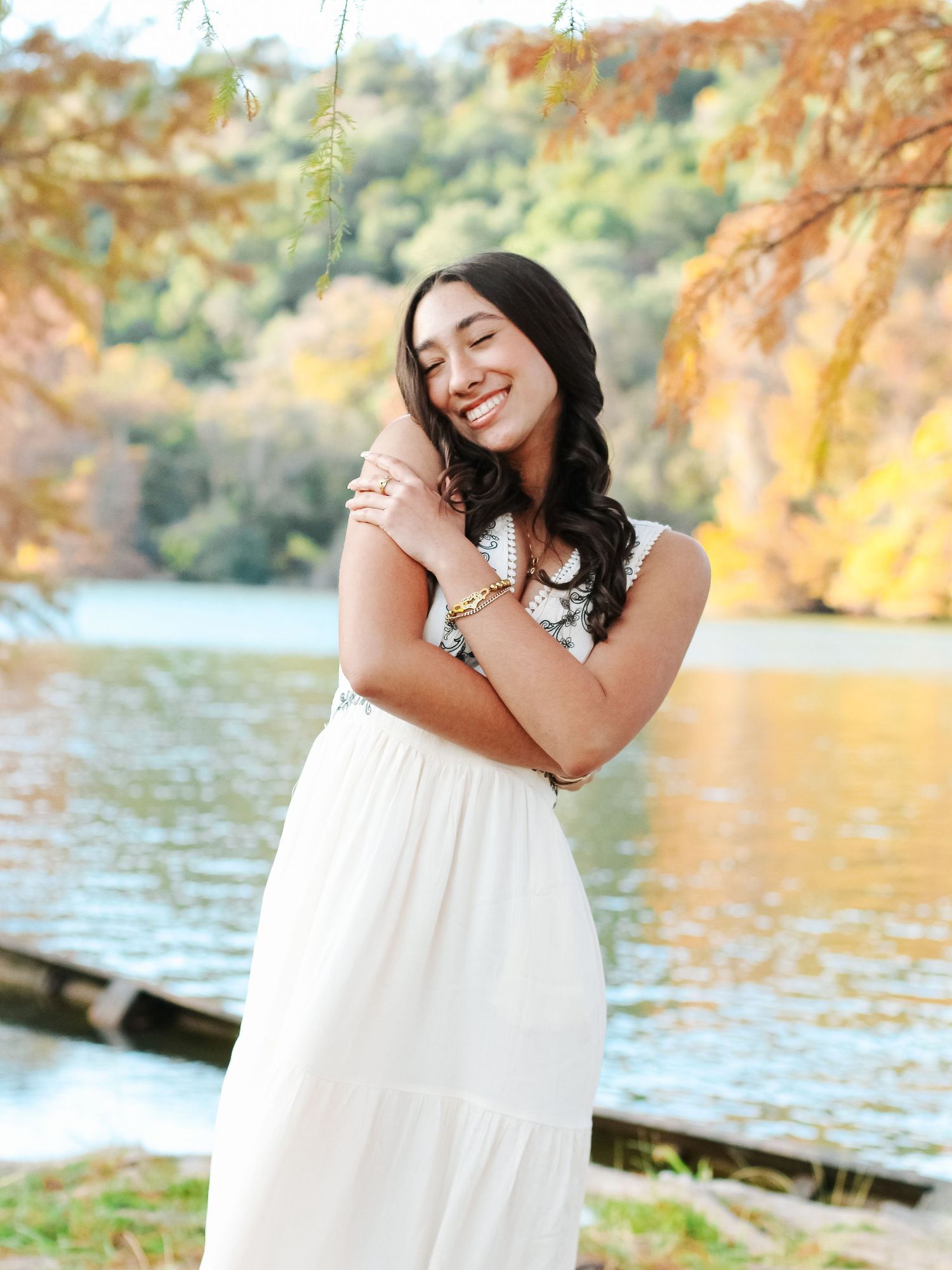 Young woman in white maxi dress smiling by autumn lake with colorful foliage backdrop.