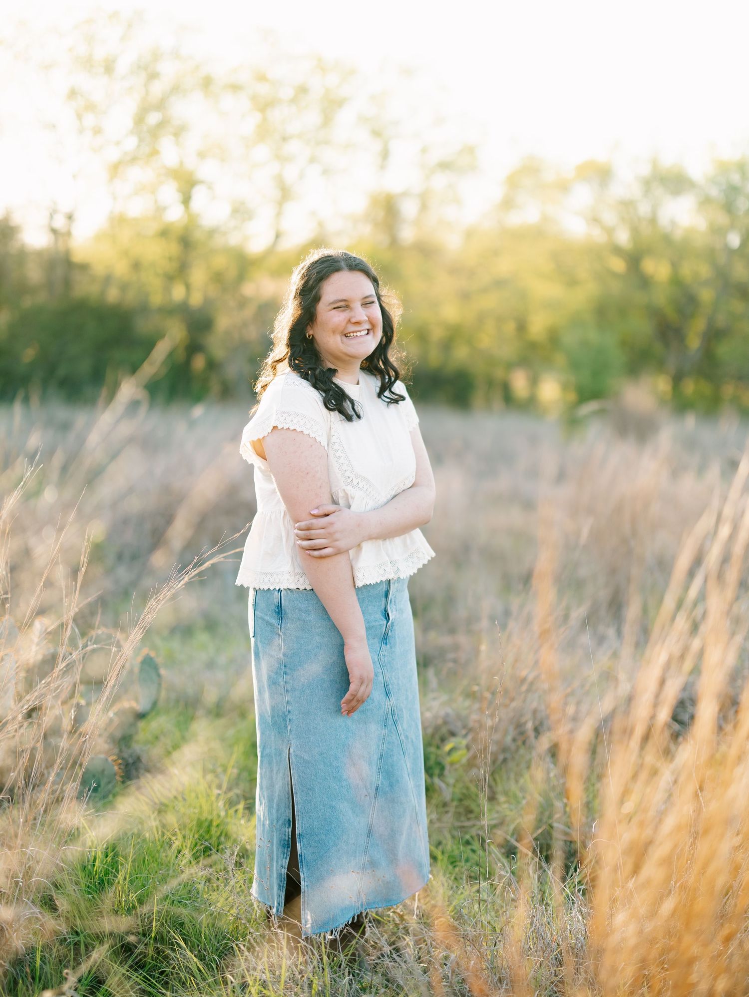 Young woman in white top and denim skirt smiling in golden hour field.