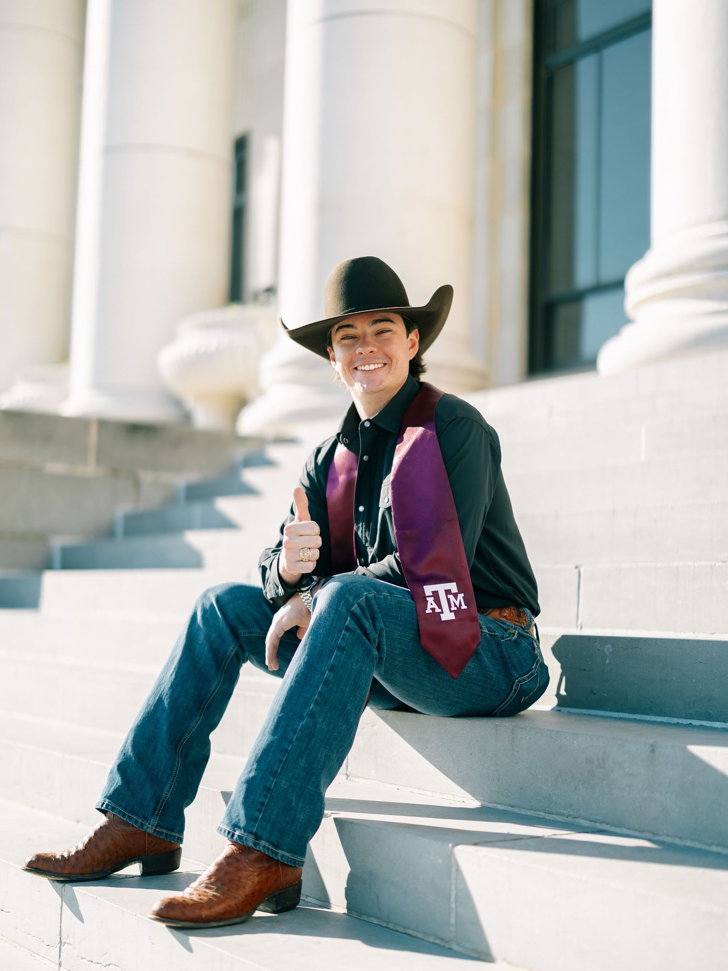 Smiling student in cowboy hat and boots with Texas A&M graduation stole sitting on stone steps.