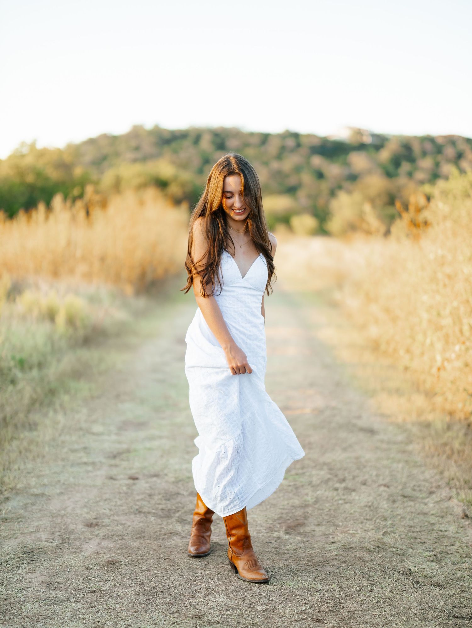 Young woman in white maxi dress and brown cowboy boots walking on a dirt path through golden fields.
