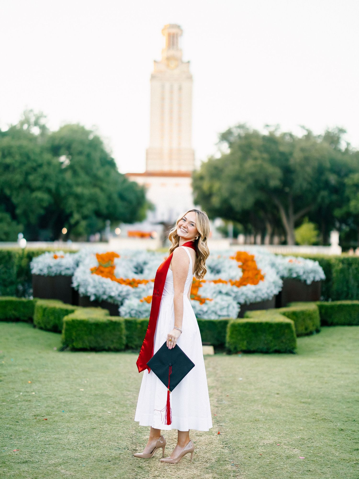 Smiling graduate in white dress holding cap near UT Austin tower with colorful flower gardens.