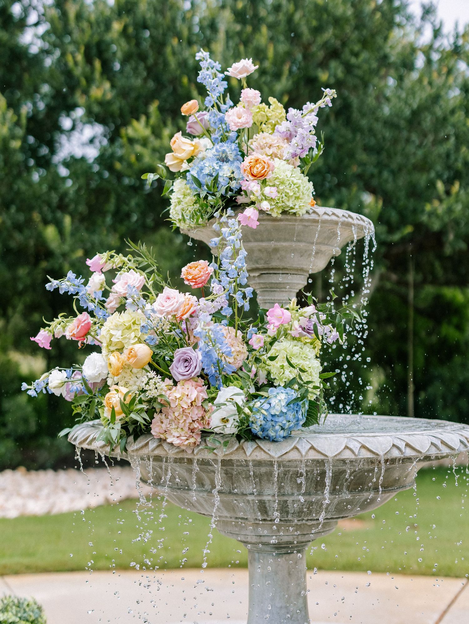 Stone tiered fountain adorned with colorful spring flowers including blue hydrangeas, pink roses, and peach blooms in a garden setting.
