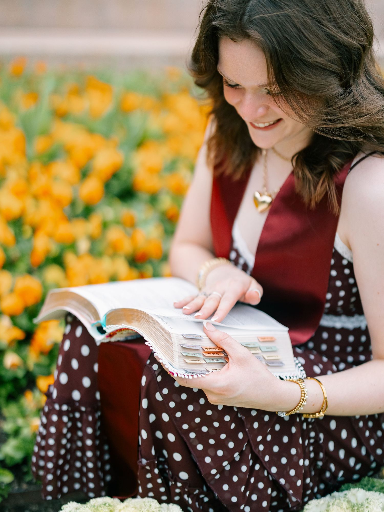 Young woman in polka dot dress reading a book outdoors among vibrant yellow tulips, smiling softly.