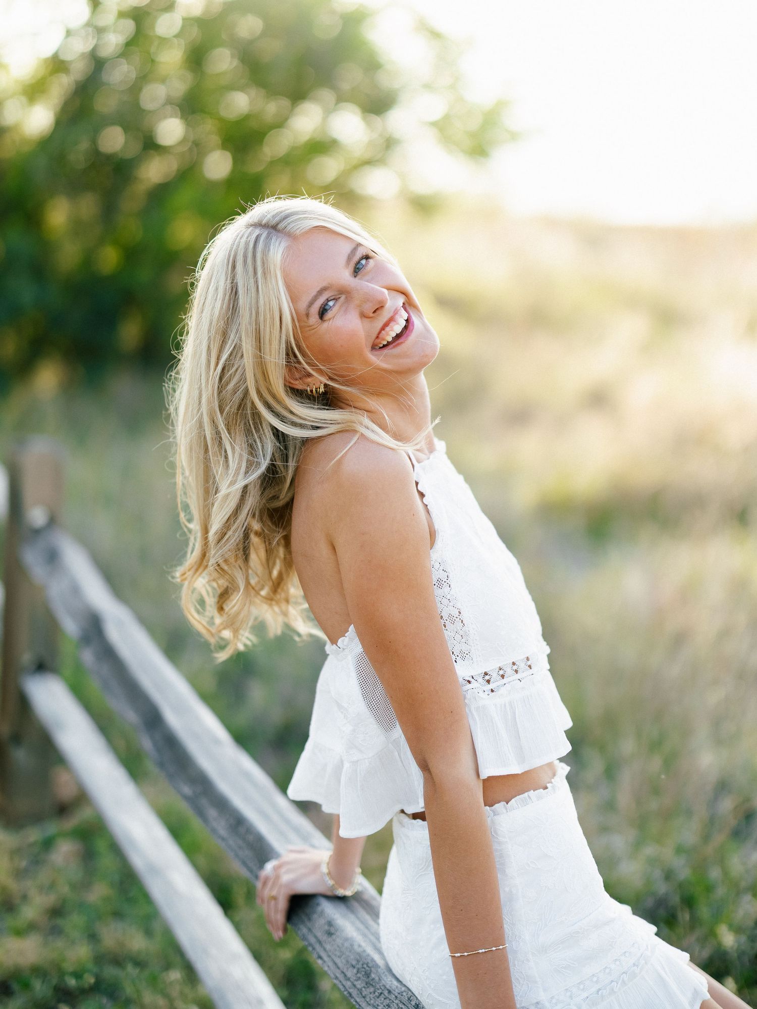 Smiling blonde woman in white crop top and skirt leaning on wooden fence in sunny outdoor field.