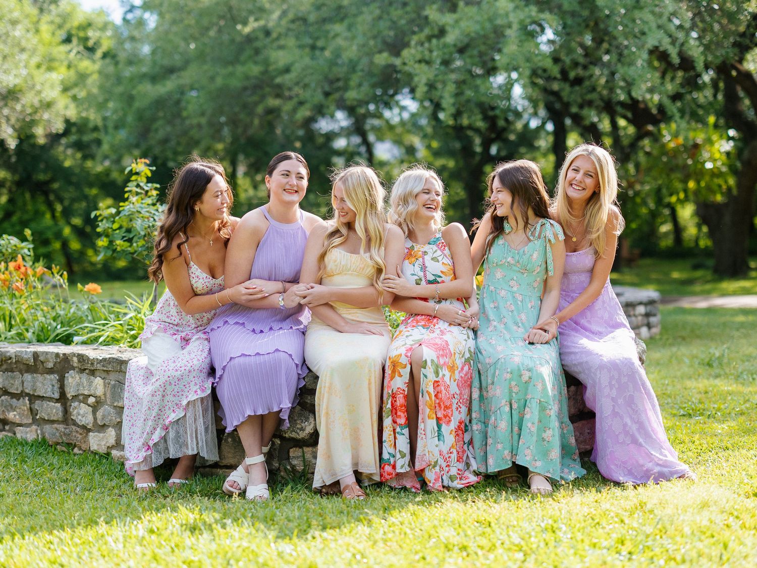 Six women in colorful spring dresses sitting together on a stone wall outdoors, laughing and smiling.