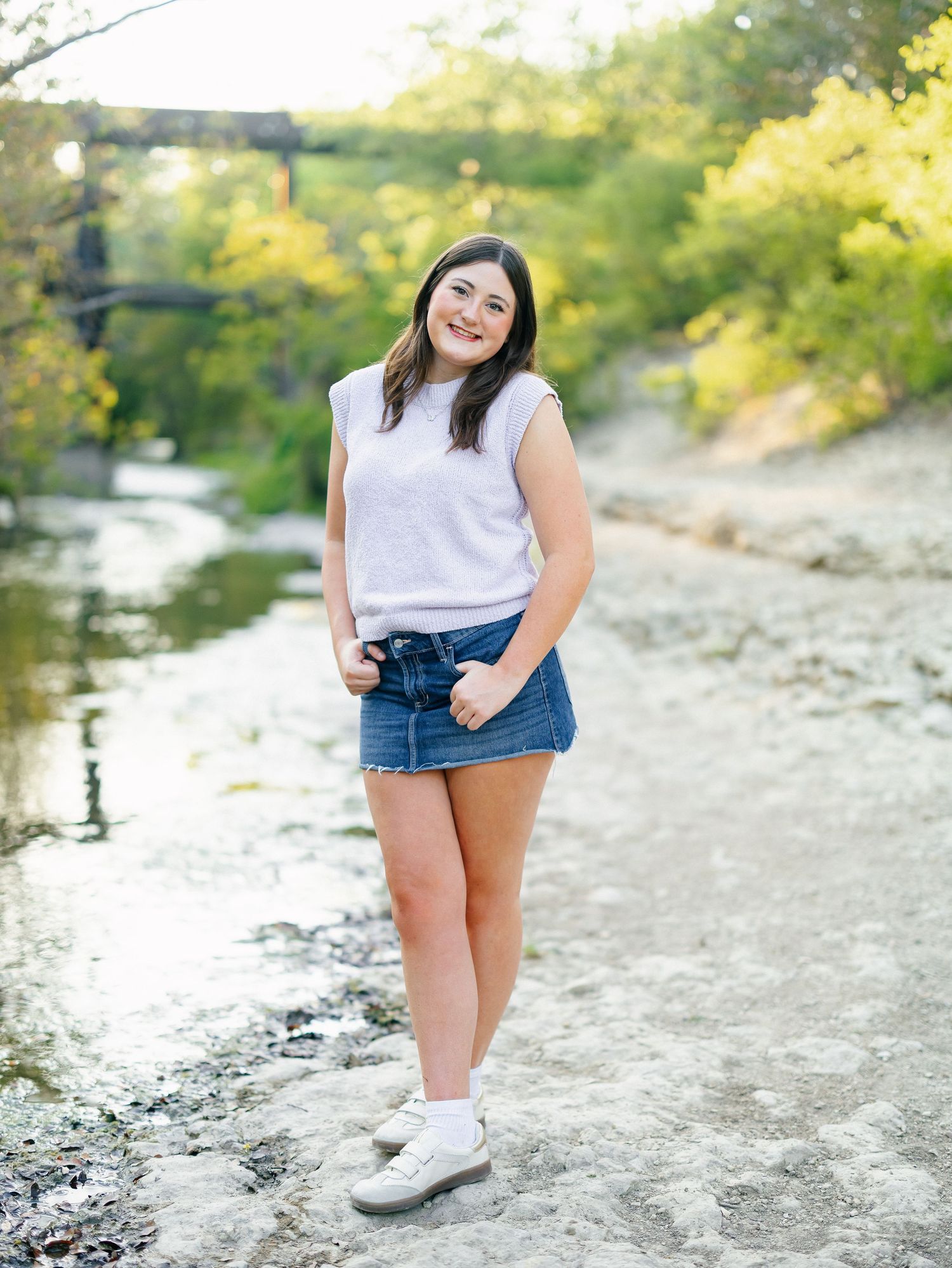 A smiling young woman in a white sleeveless top and denim mini skirt poses by a rocky creek with lush green trees.