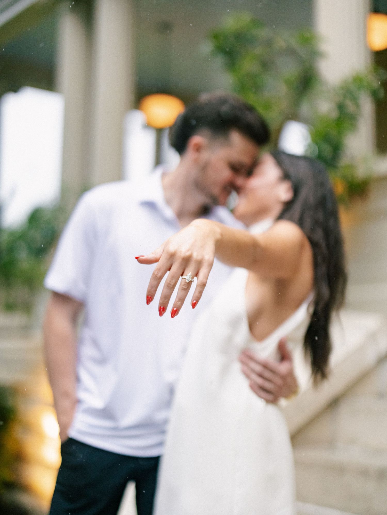 Engaged couple kissing while bride shows off diamond engagement ring with red nails in romantic outdoor portrait.