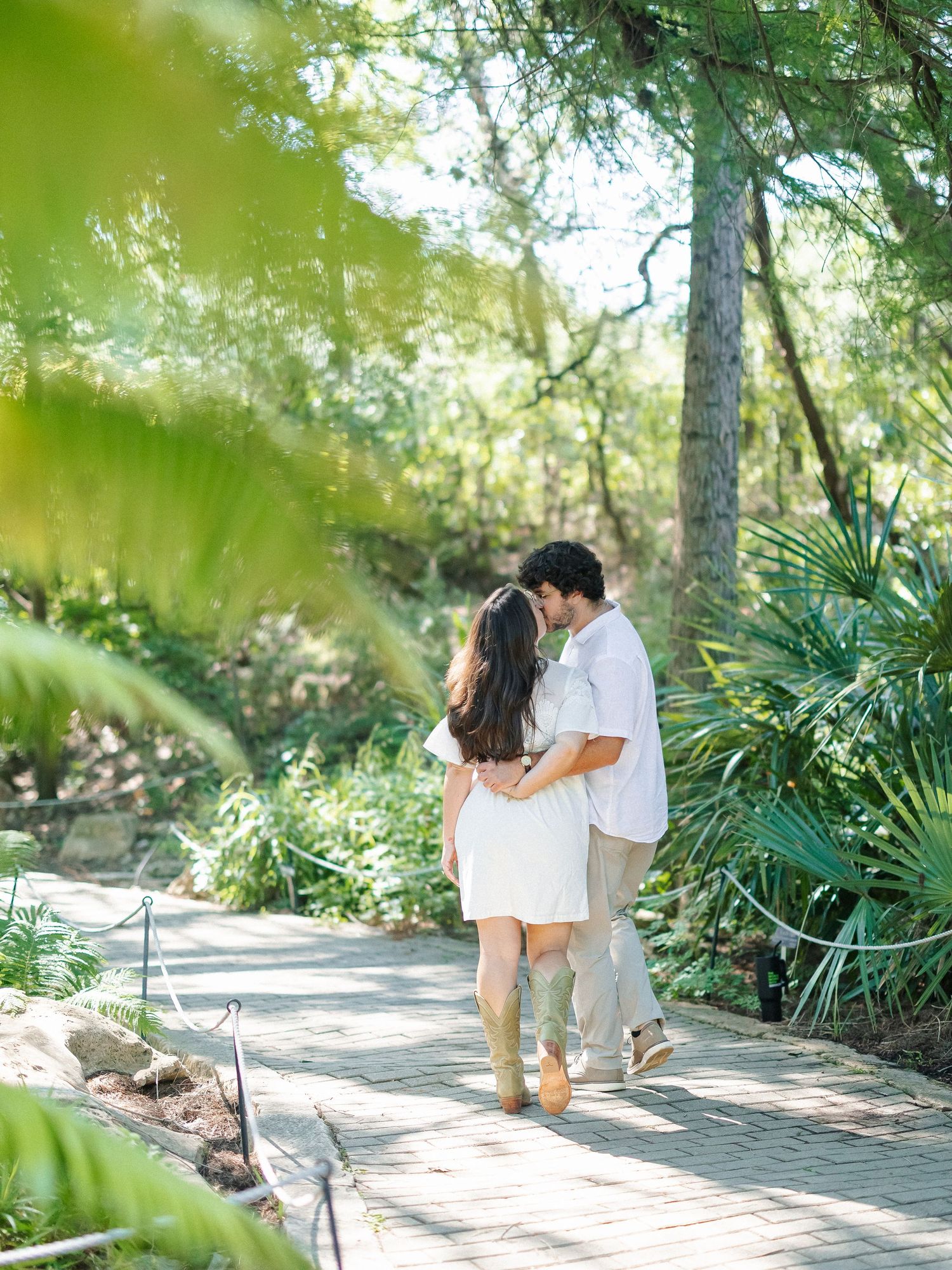 Couple walking hand-in-hand on a garden path surrounded by lush green trees and tropical plants.