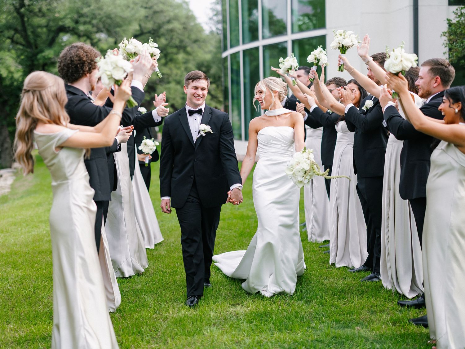 Bride and groom walk through cheering wedding party in black and white attire on green lawn.