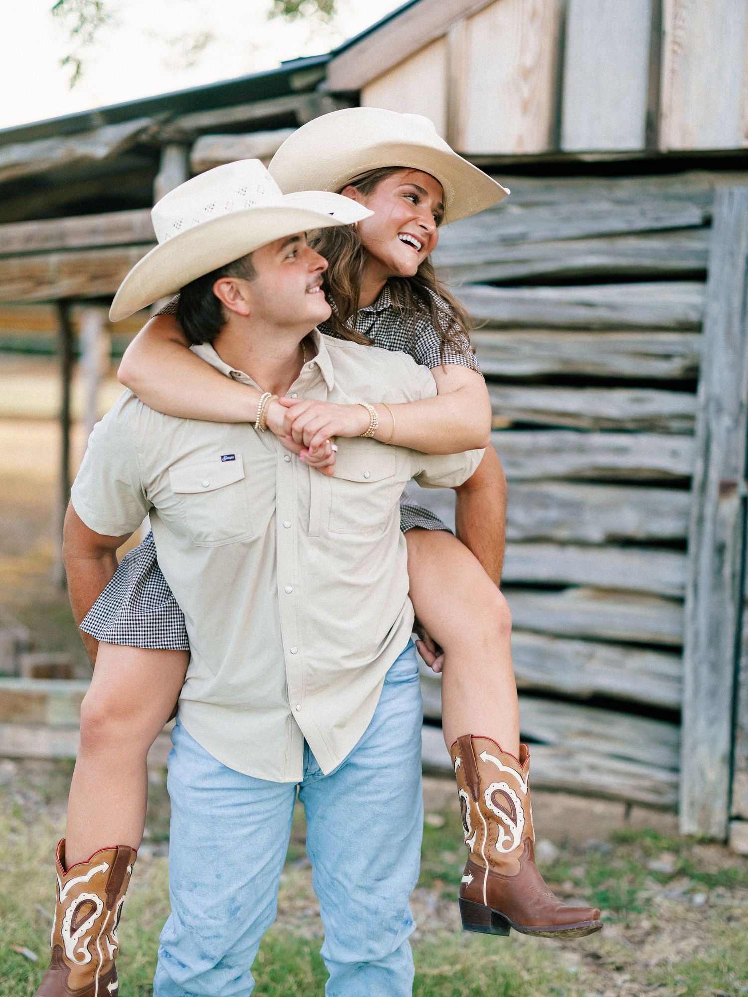 Smiling couple in matching cowboy hats posing playfully with woman on mans back near rustic log structure.