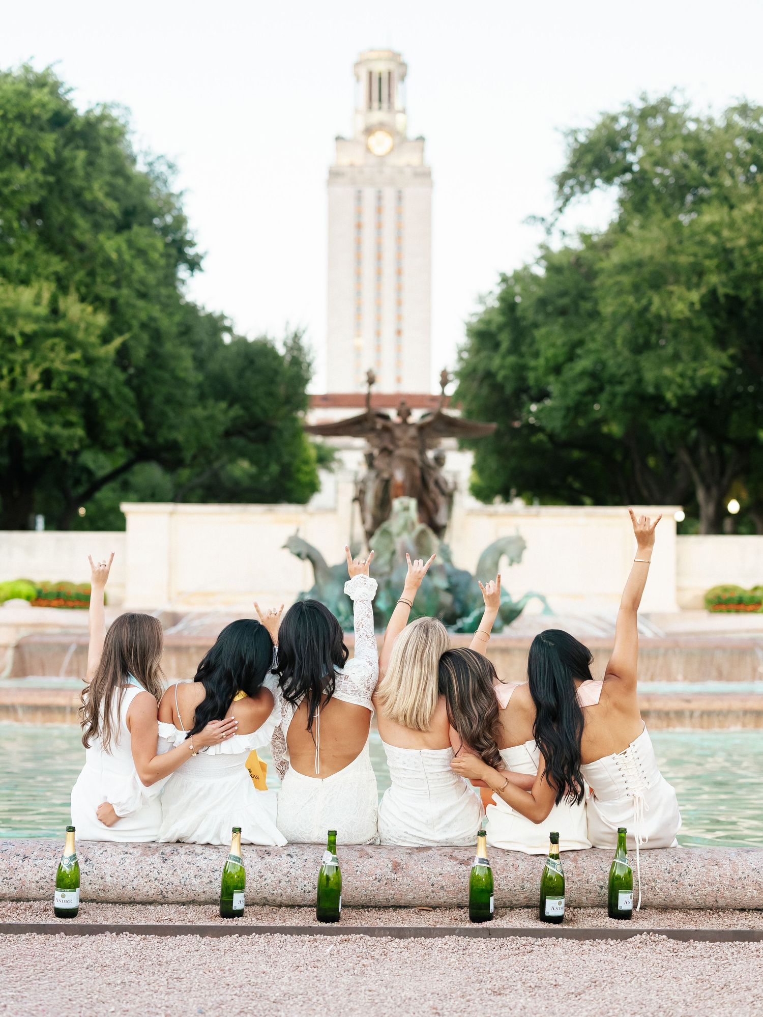 Five women in white sit by a fountain raising champagne bottles at the University of Texas campus.