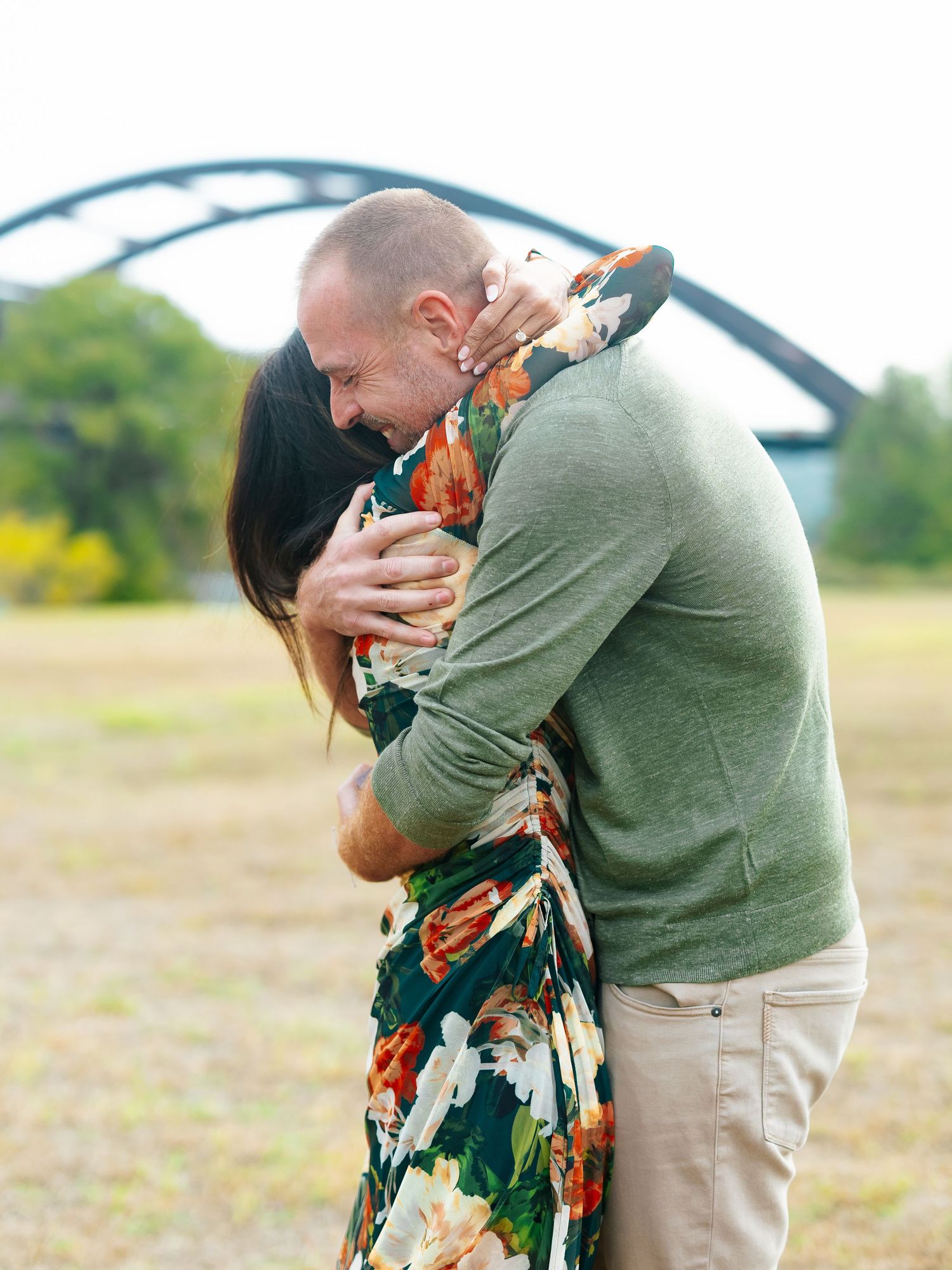Couple sharing a warm embrace in an open field with an iconic arched bridge in the background.
