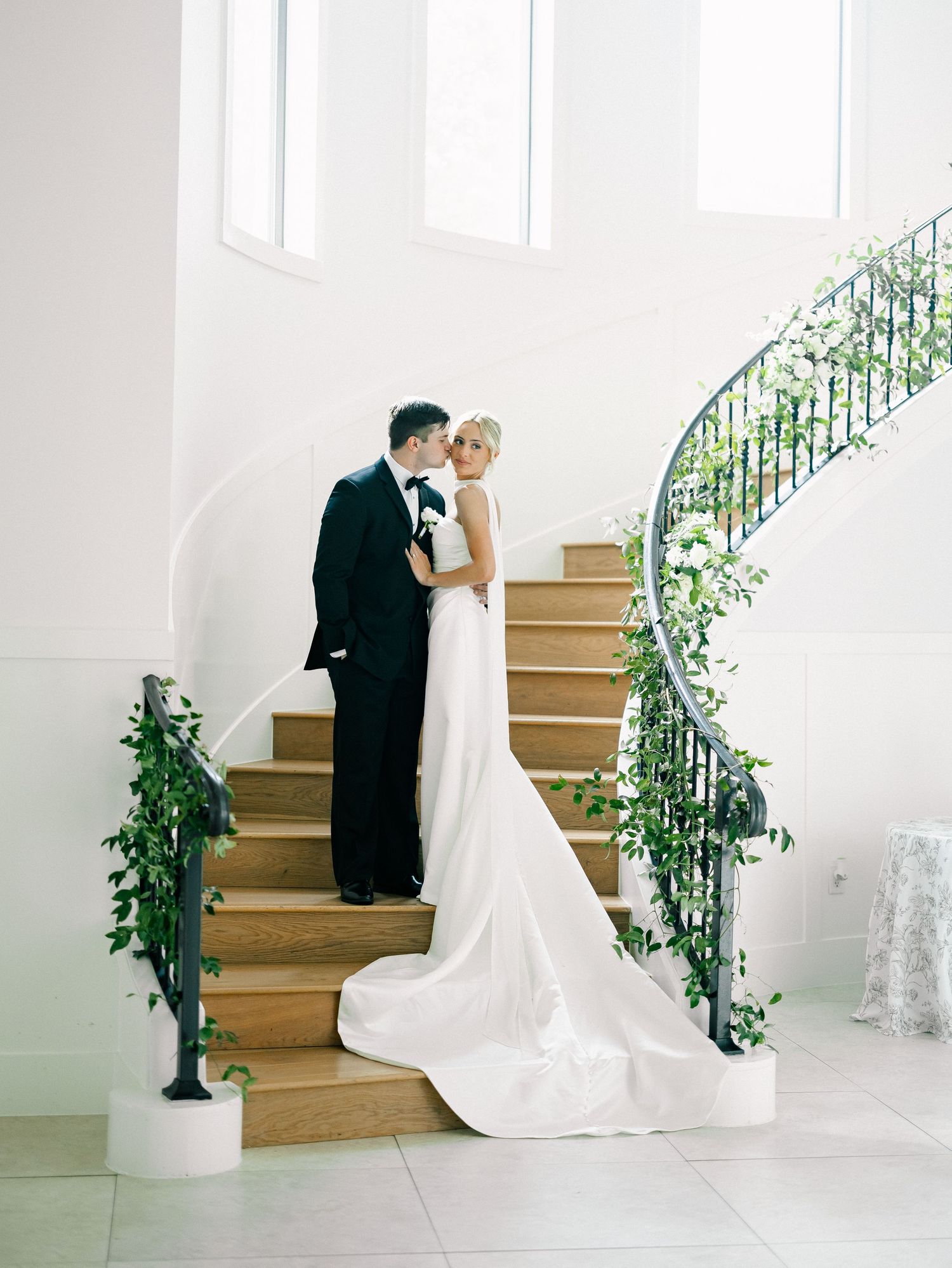 Bride and groom share a kiss on elegant spiral staircase adorned with lush greenery in a bright white wedding venue.