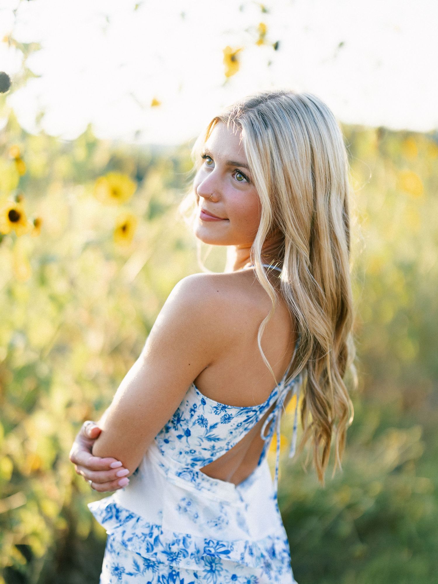 Blonde girl in blue floral dress poses among sunflowers in golden sunlight.
