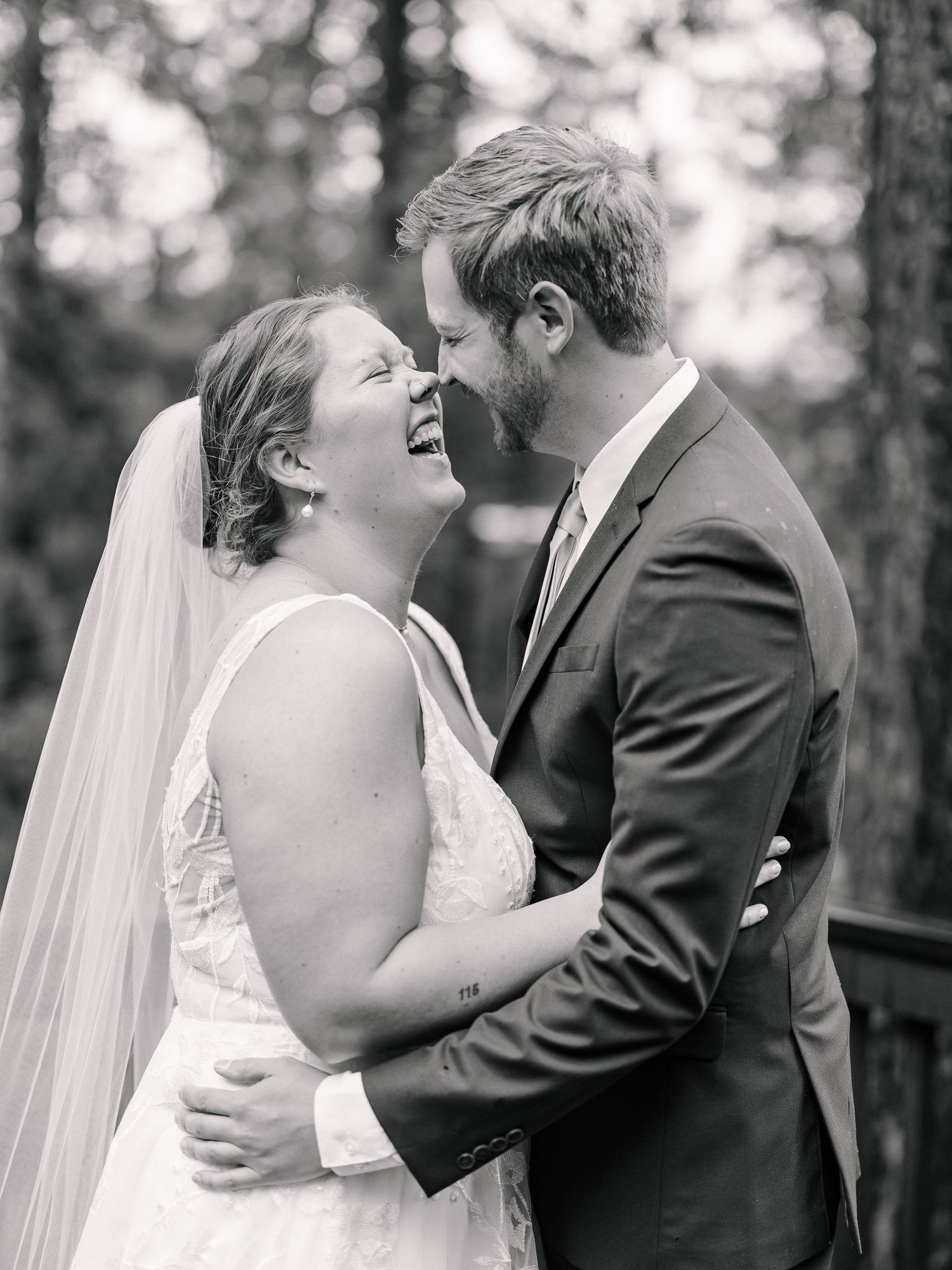 Black and white photo of laughing bride and groom sharing an intimate moment outdoors among trees.