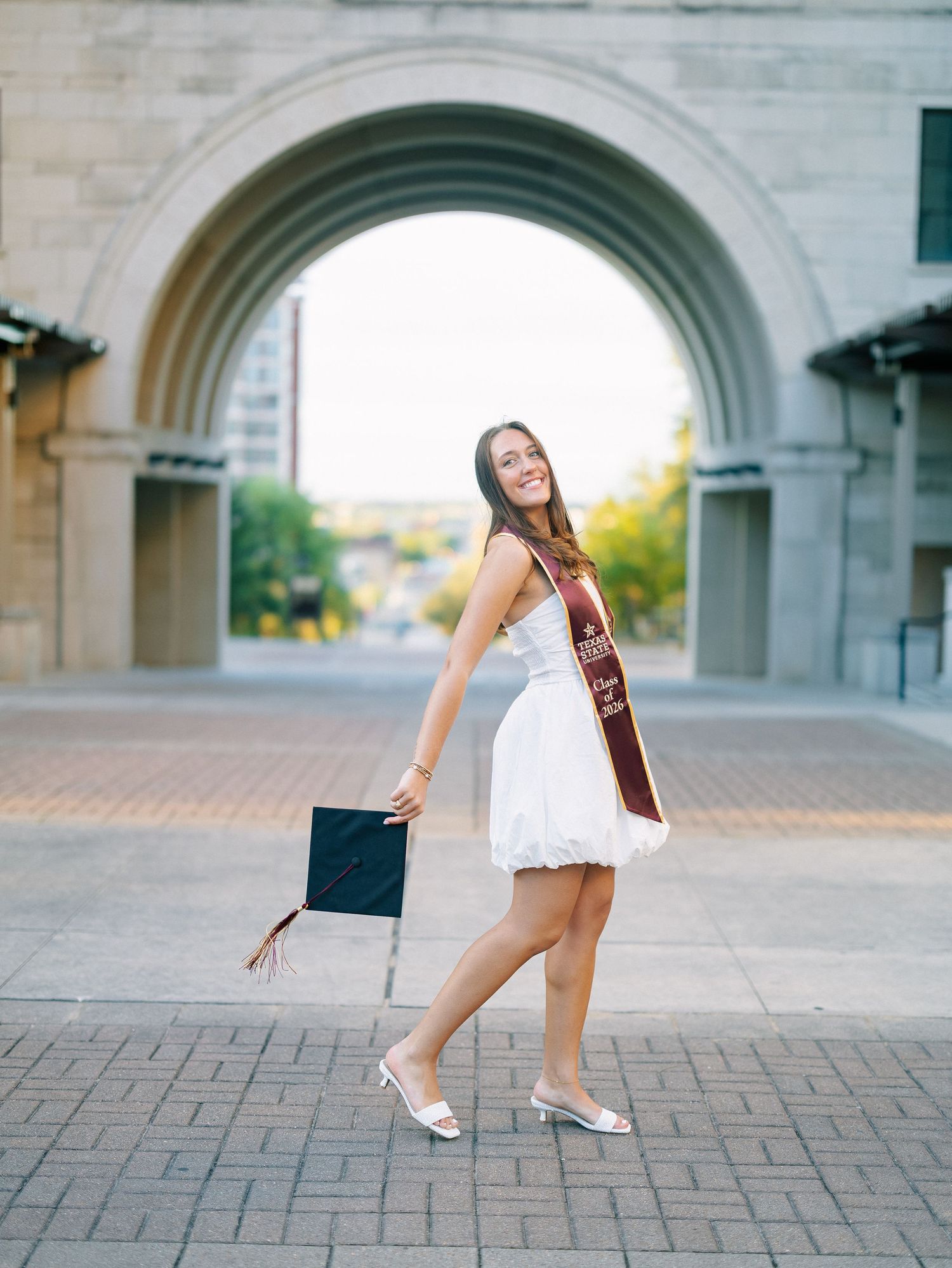 Woman in white dress holding graduation cap while posing under a stone arch on campus.
