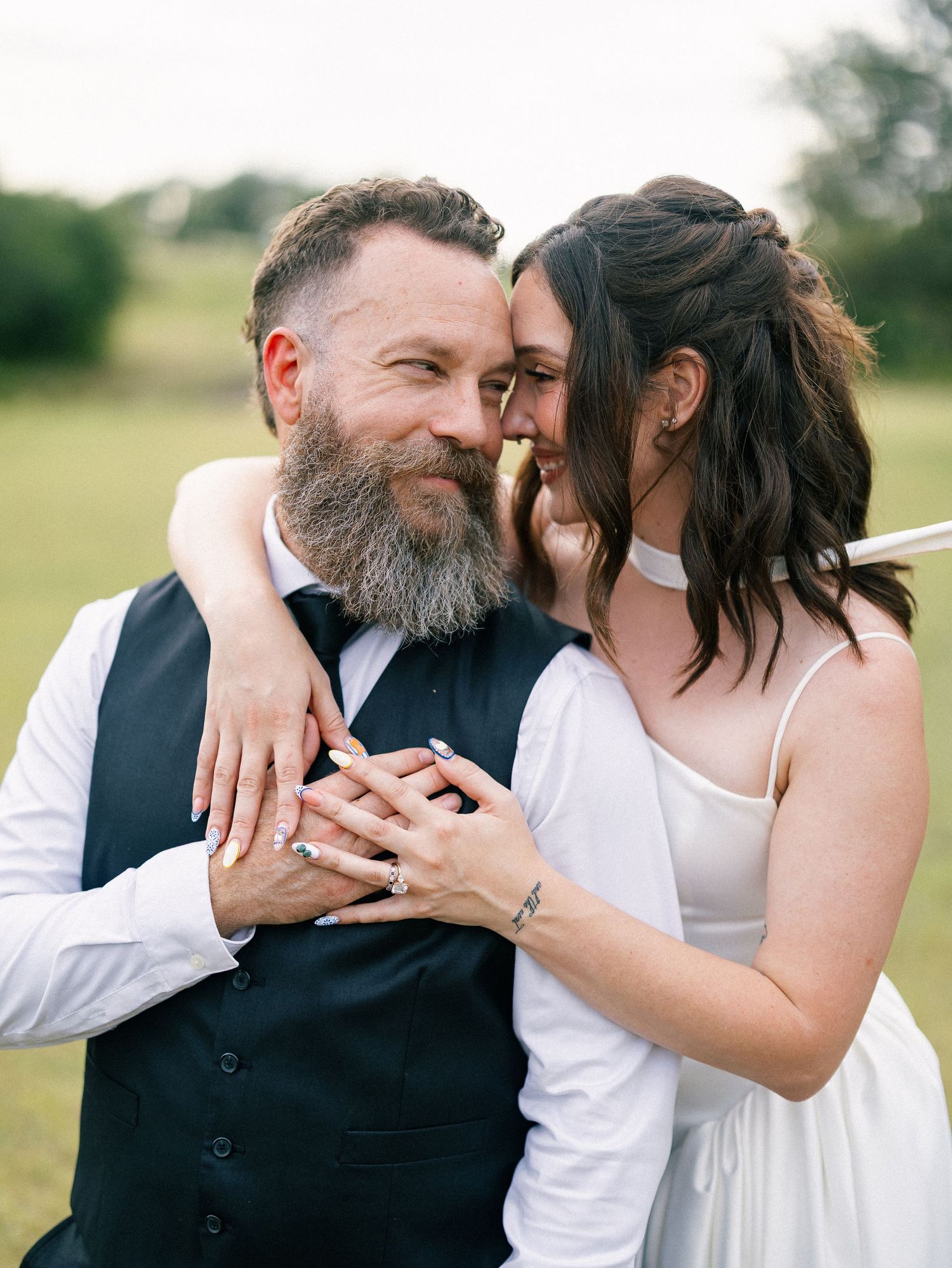 A bearded groom in a vest smiles as his bride in a white dress leans close, showing off wedding rings outdoors.