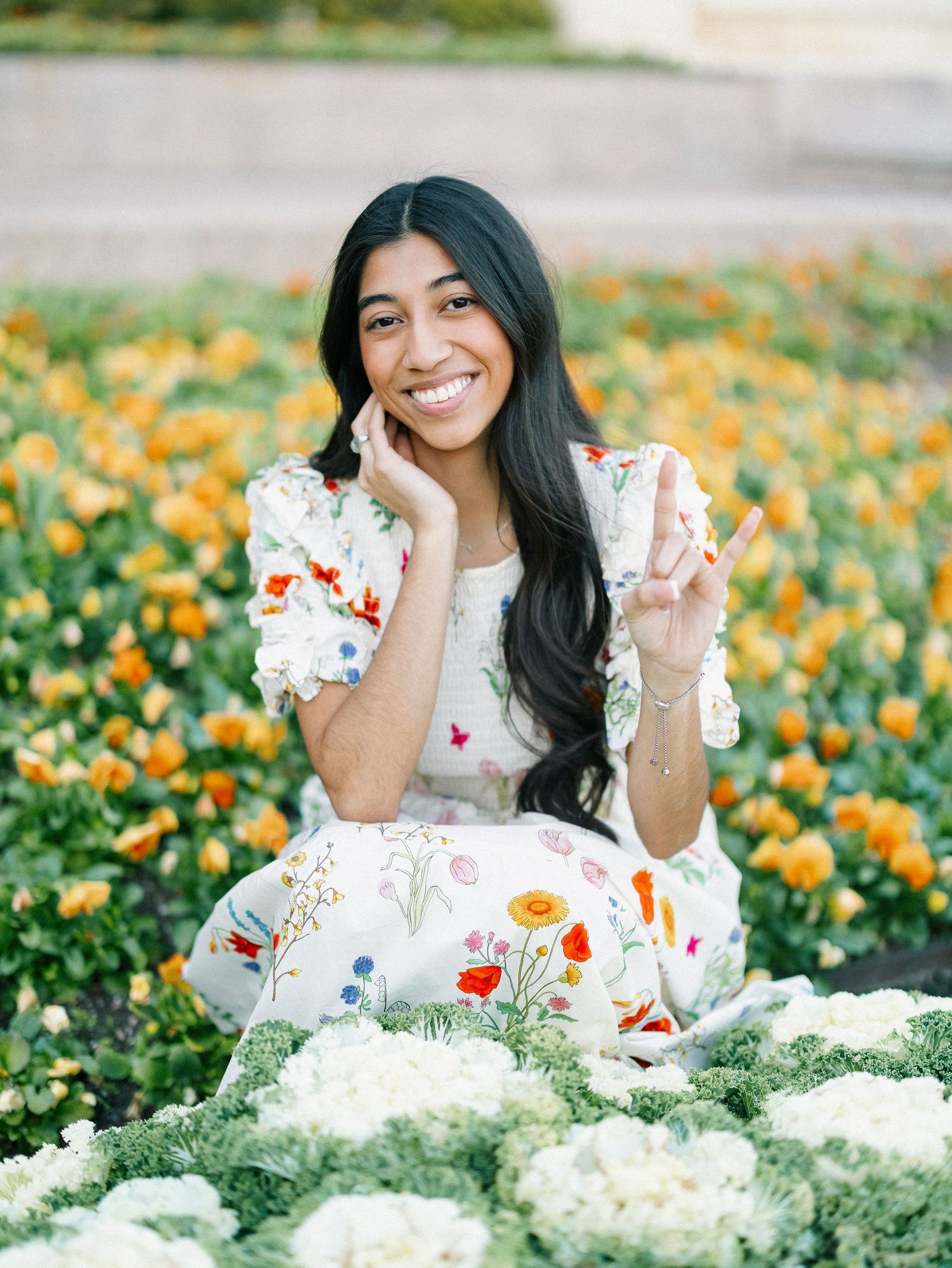A smiling young woman in a floral dress sits among colorful blooming flowers in a garden.