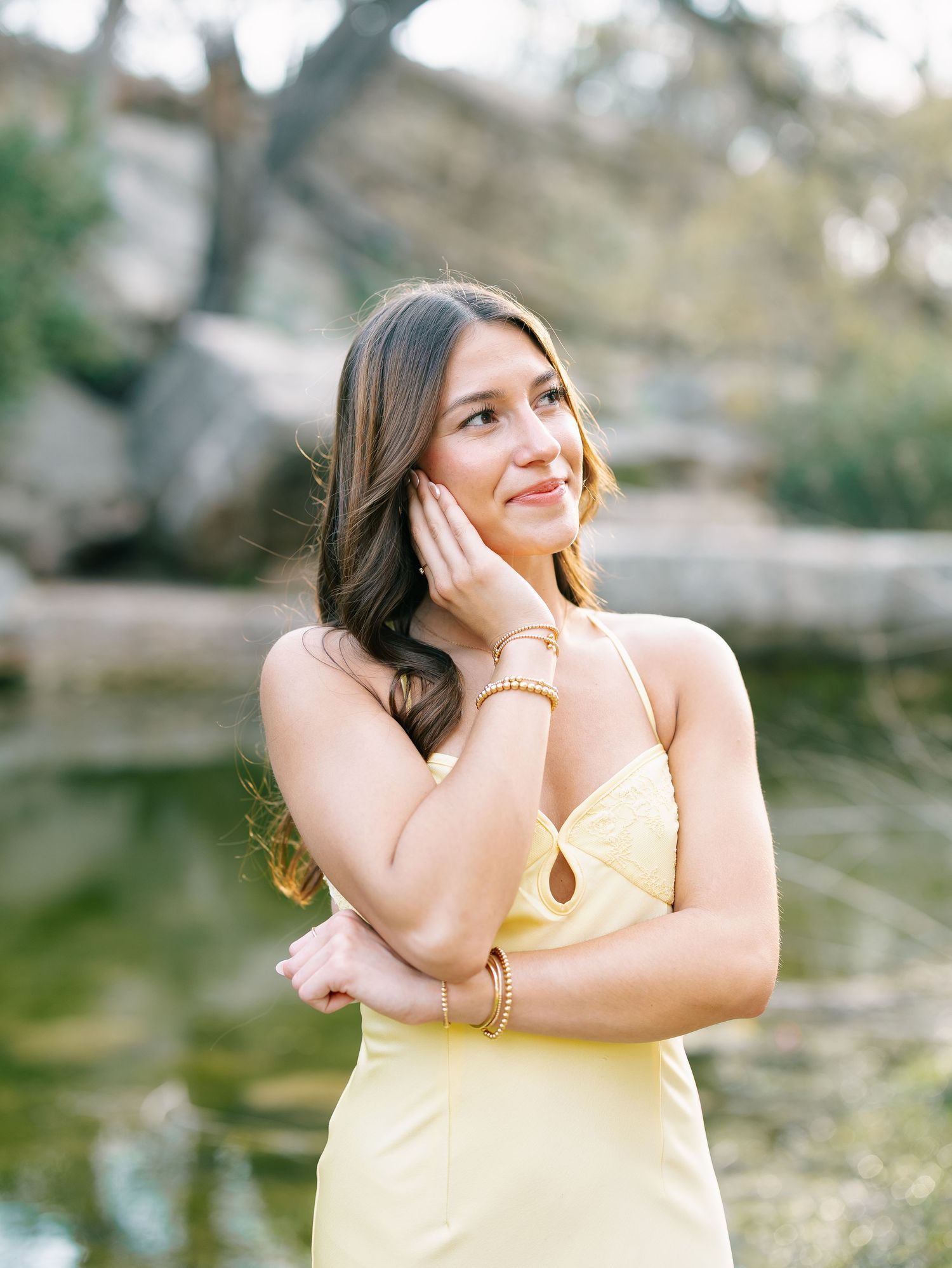 Young woman in yellow halter dress poses outdoors near rocky creek, smiling softly with hand near face.