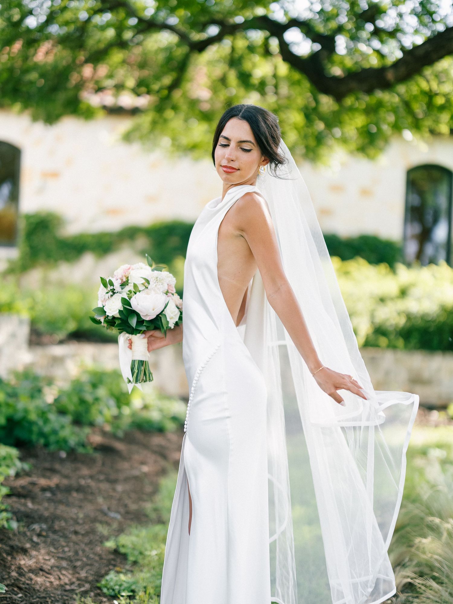 Bride in a sleek white backless gown holding a white bouquet, wearing a flowing veil outdoors near a stone building.