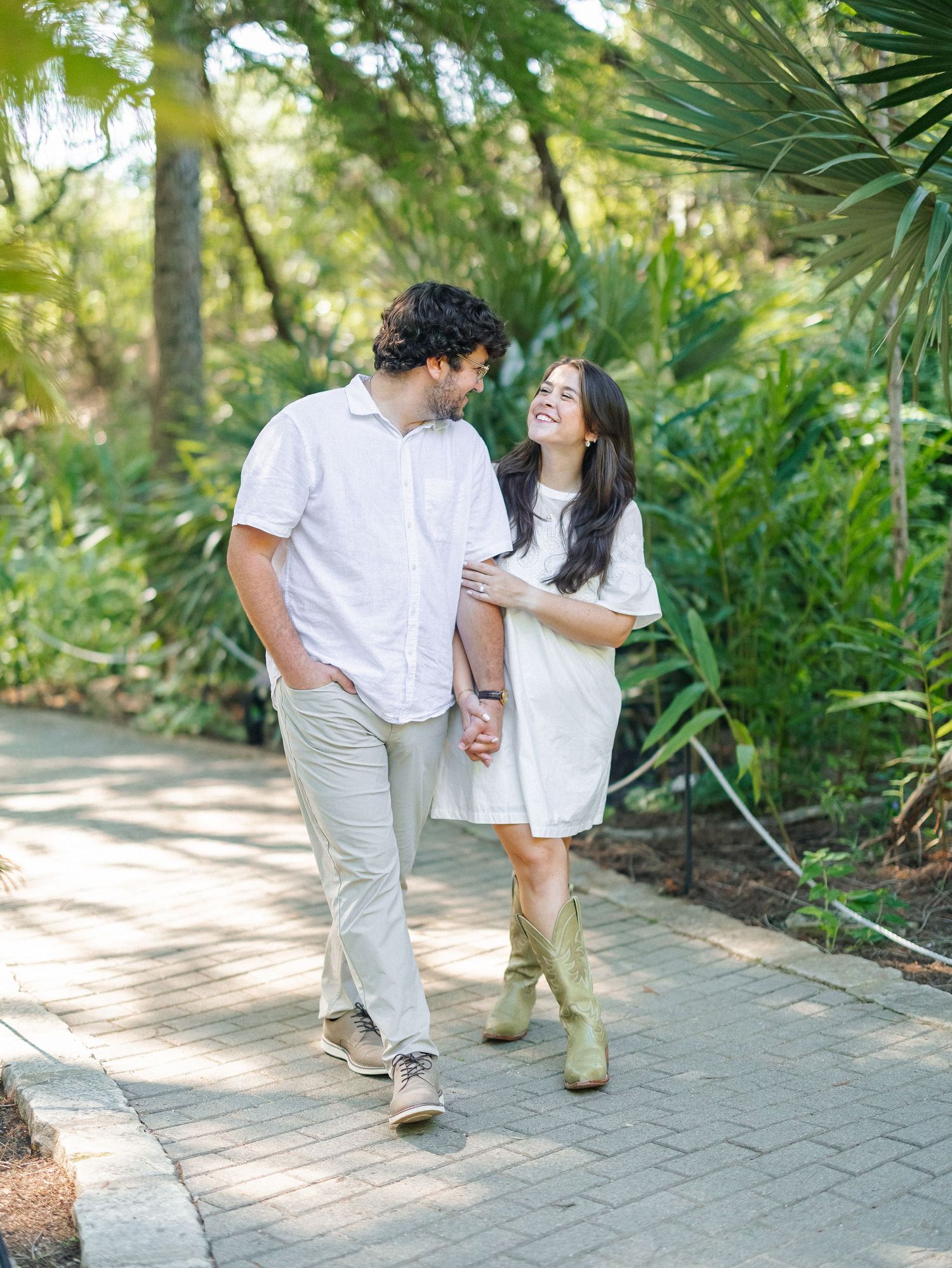A couple walks hand-in-hand on a garden path, smiling at each other, surrounded by lush green tropical plants.