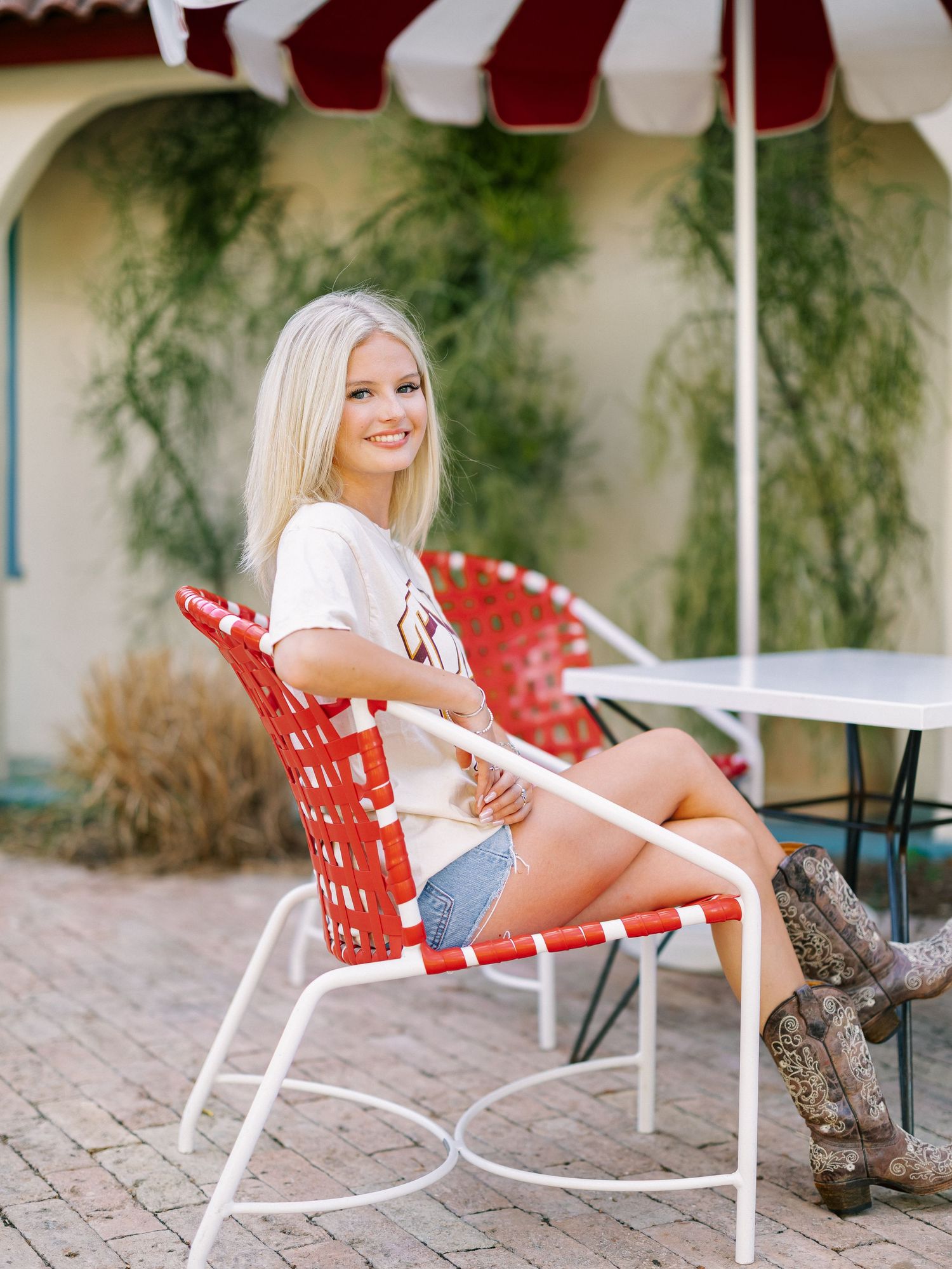 Blonde woman in denim shorts and cowboy boots sitting in a red patio chair under a striped umbrella outdoors.