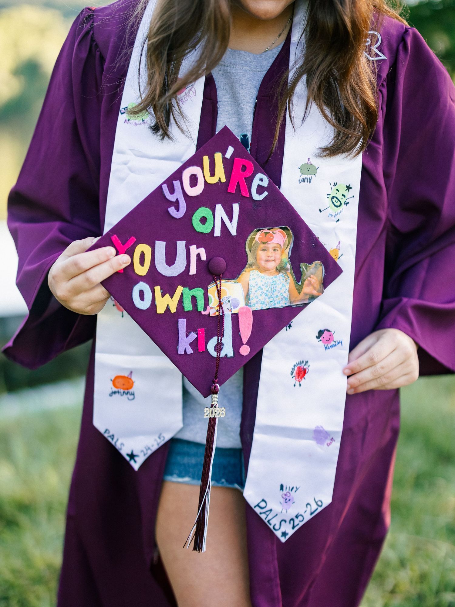 Graduate in purple gown holding decorated mortarboard cap reading you're on your own kid with childhood photo.