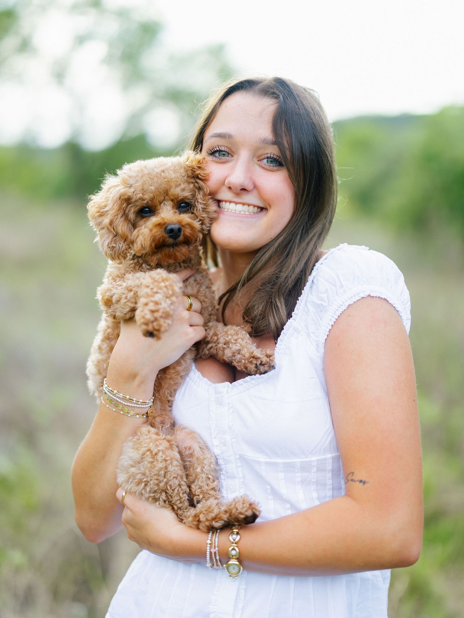Smiling young woman in white dress holding a fluffy brown toy poodle puppy outdoors in a green field.