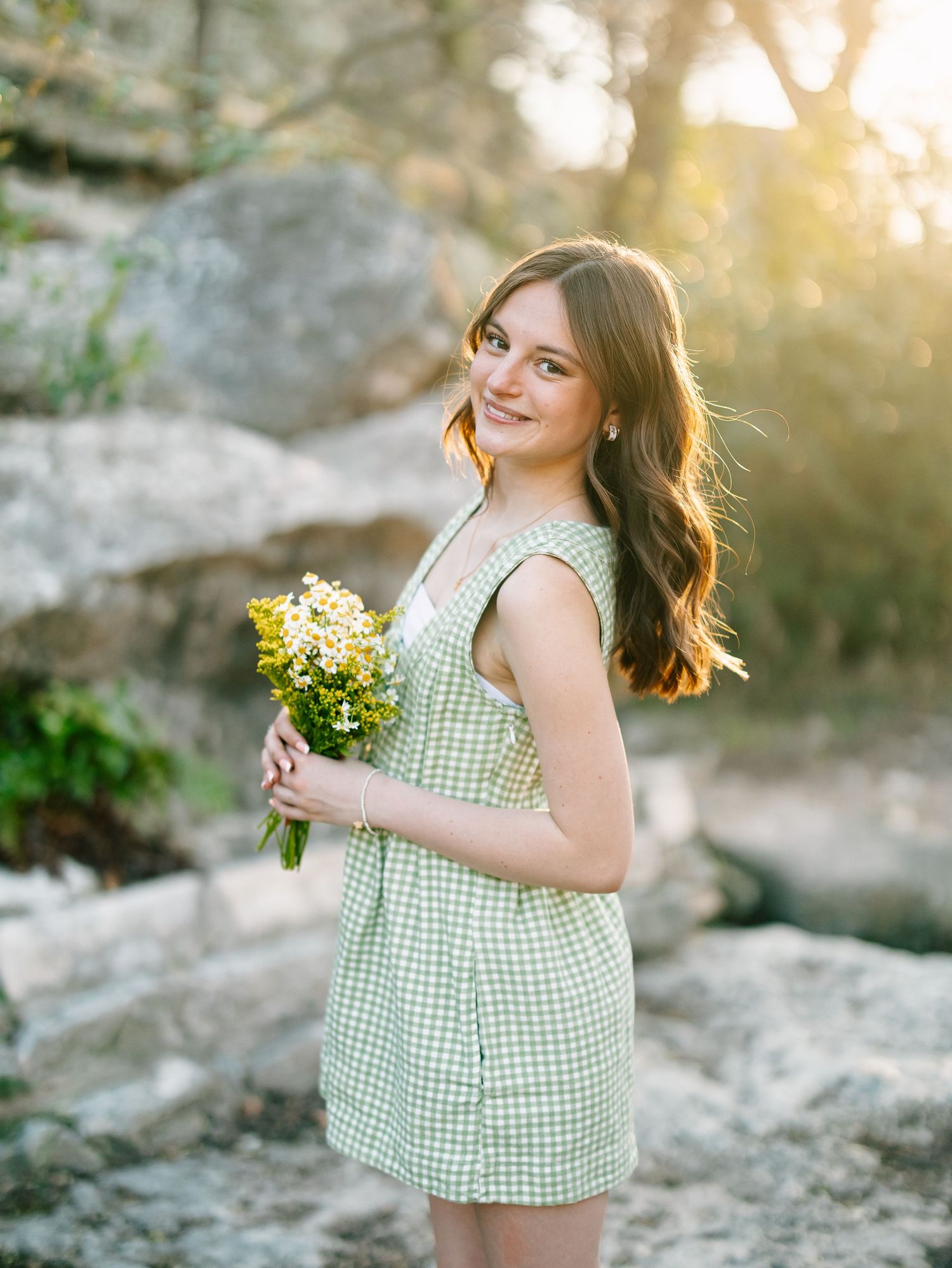 Young woman in green gingham dress holding wildflowers, smiling outdoors with golden sunlight and rocks in background.
