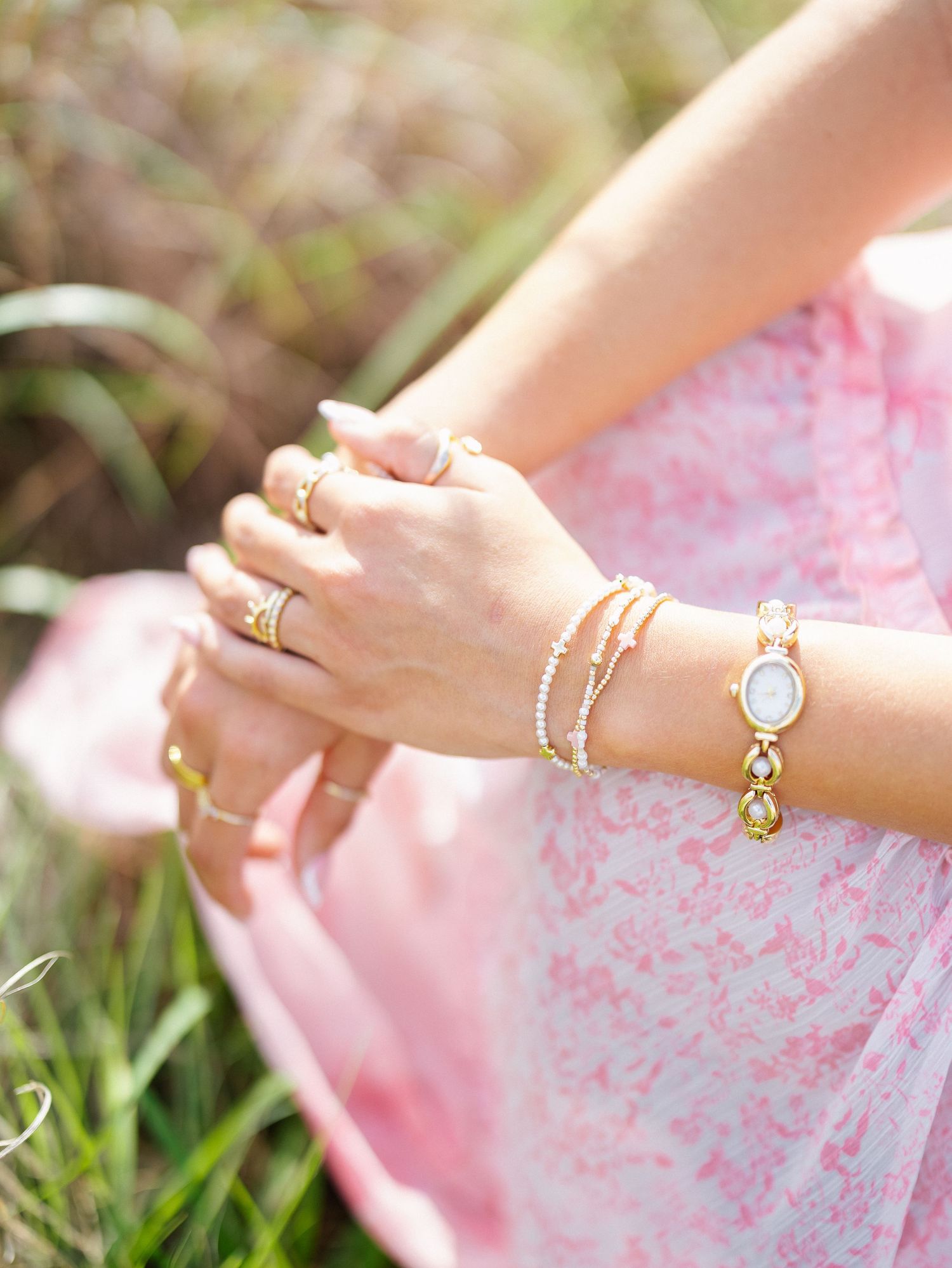 Woman wearing gold bracelets, watch, and rings with a pink floral dress, sitting outdoors in a garden setting.