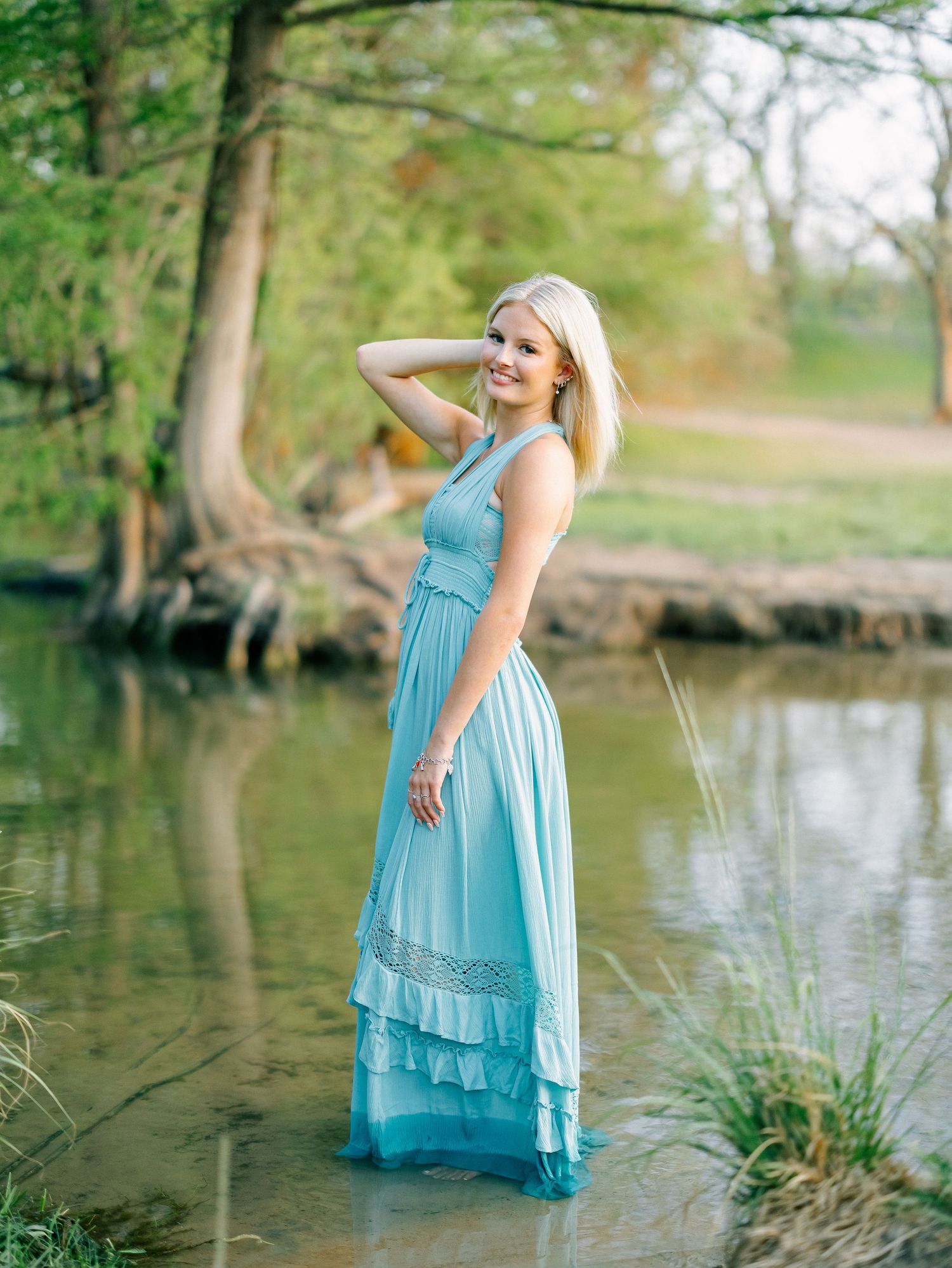Blonde woman in teal maxi dress standing in shallow creek surrounded by trees.