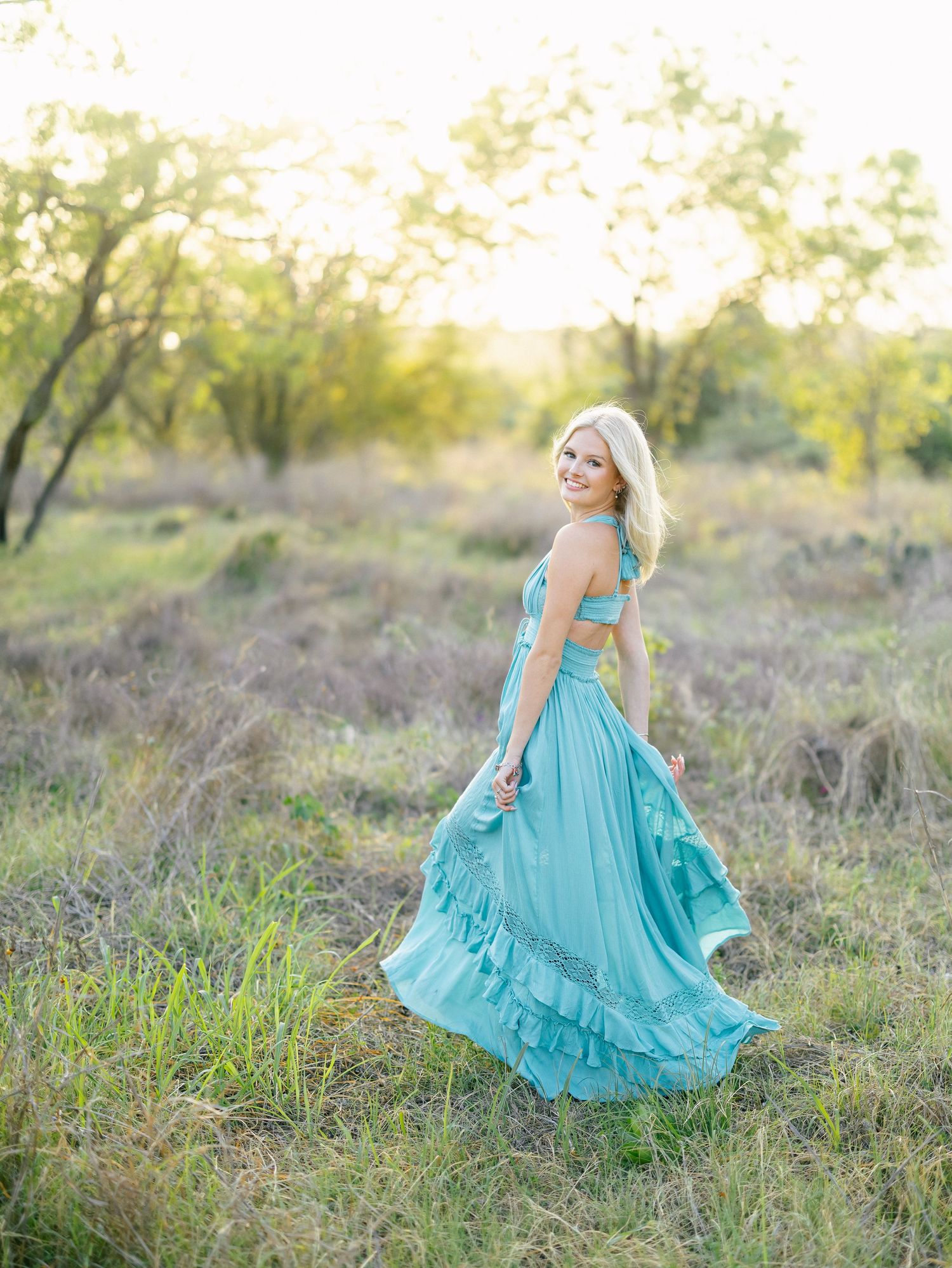 A blonde woman twirls in a flowing turquoise ruffled maxi dress in a sunlit meadow with trees in the background.