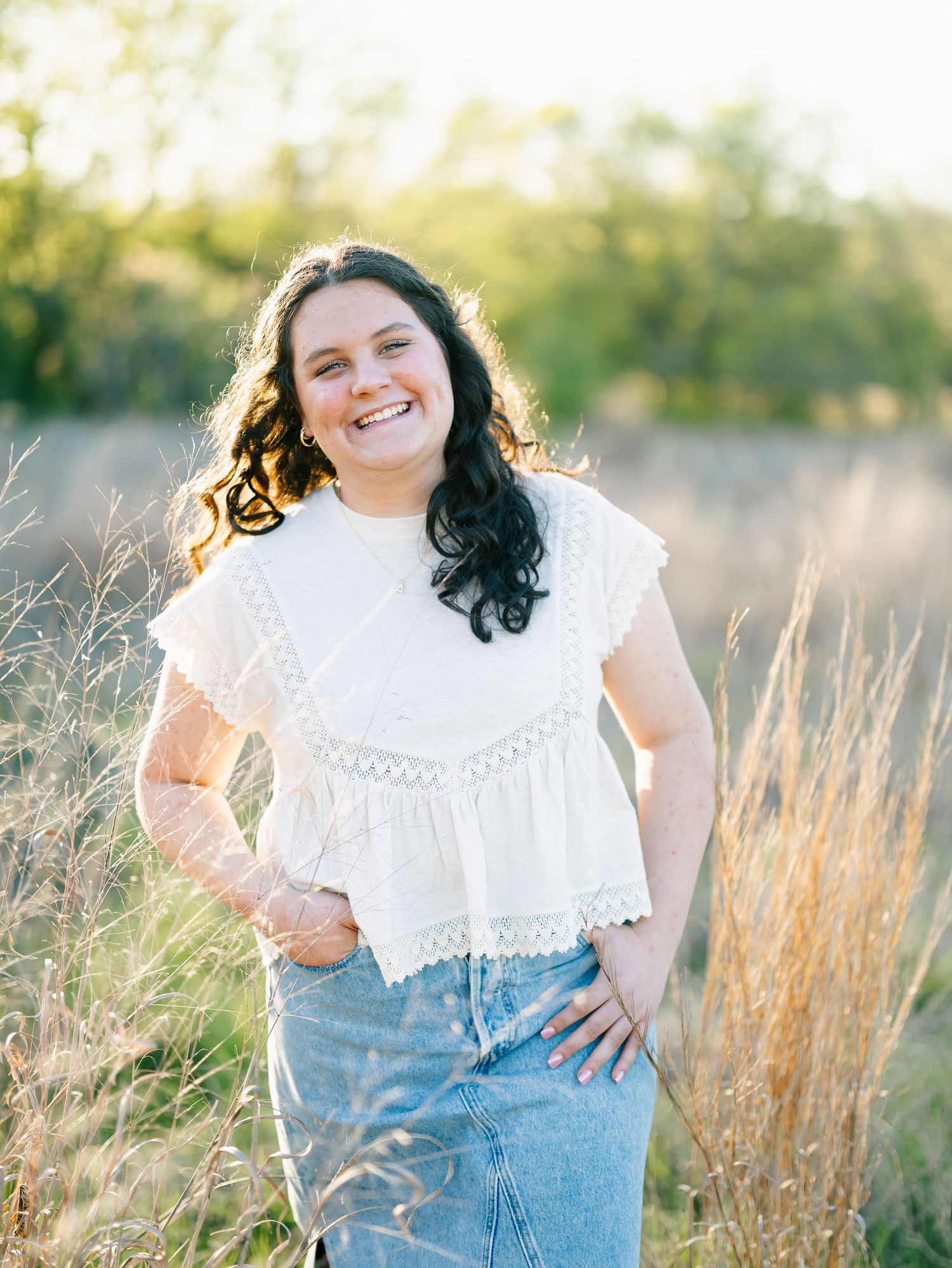Smiling young woman in white lace top and denim skirt posing in golden field at sunset.