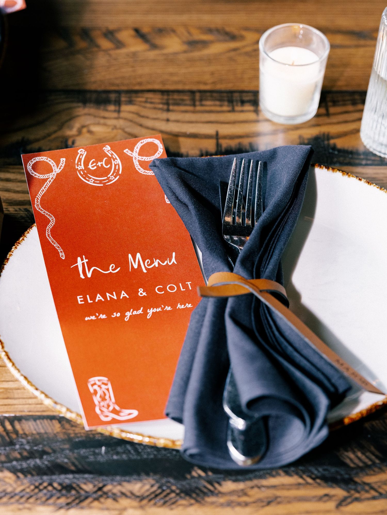 Western wedding place setting with red menu card for Elana & Colt, navy napkin, and silverware on white plate.