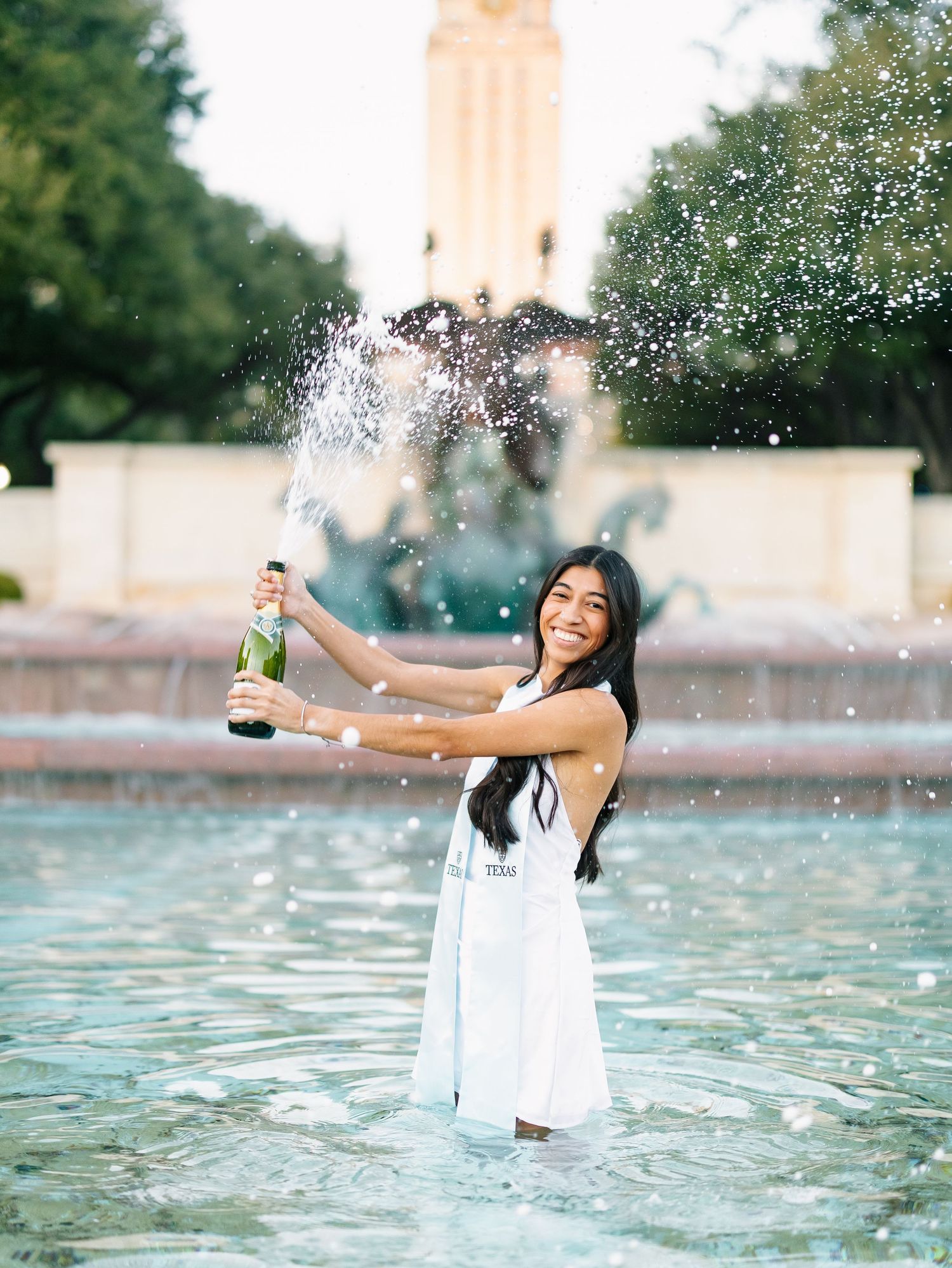 Graduate in white dress popping champagne bottle in university fountain.